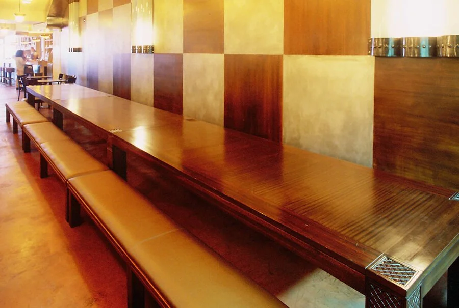Empty restaurant with a long wooden table and beige cushioned benches along the side, wooden wall panels, and some people in the background.


