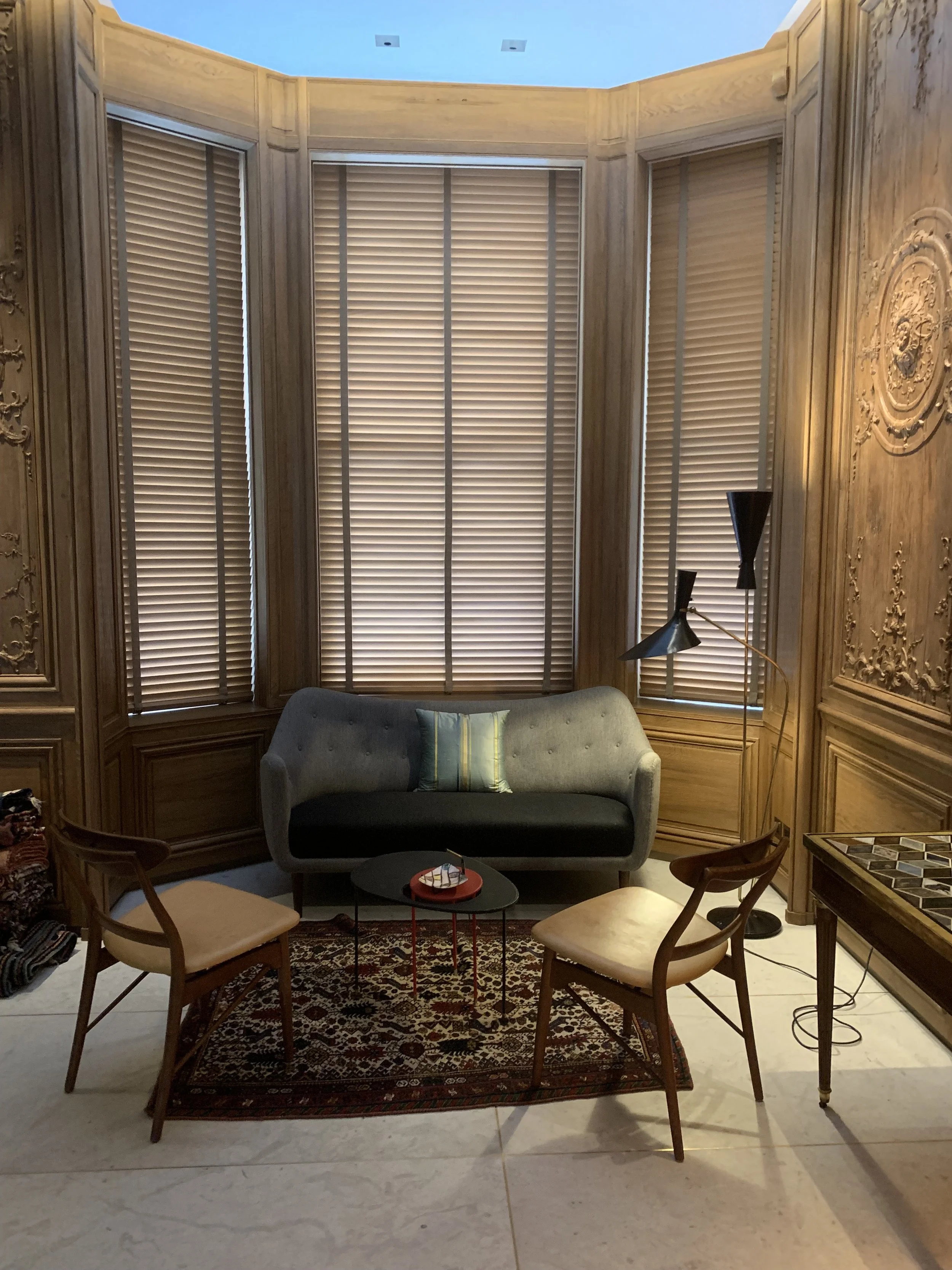 A cozy sitting area with a gray sofa, two wooden chairs with beige cushions, a small round black coffee table, and a colorful rug on a tiled floor. The room has tall wooden panelled walls with decorative carvings and large windows with closed blinds,