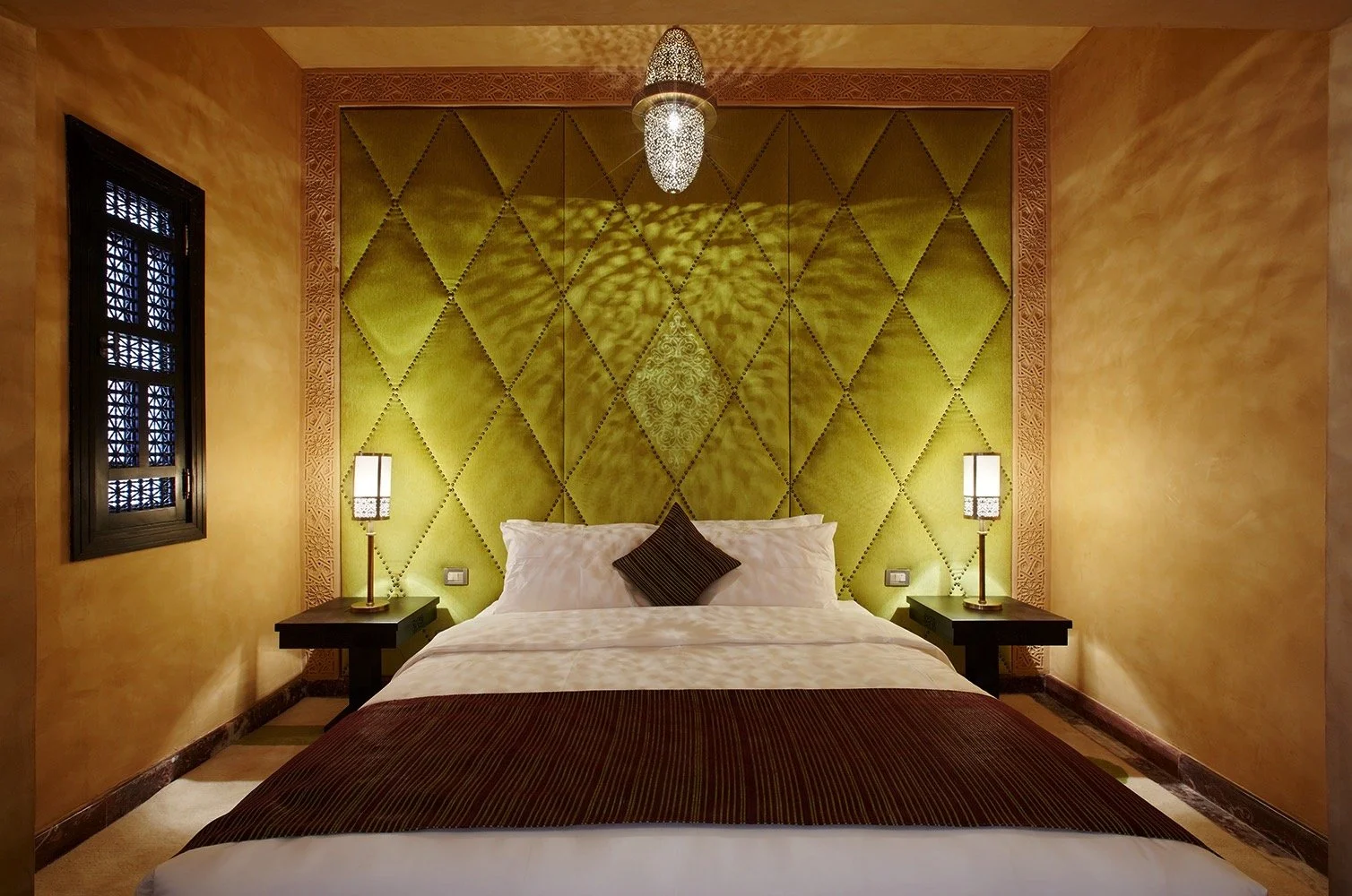 A bedroom with a green padded headboard, two black nightstands with lamps, a white bed with a dark brown pillow, and a textured yellow wall with a window on the left and a decorative light fixture hanging from the ceiling.


