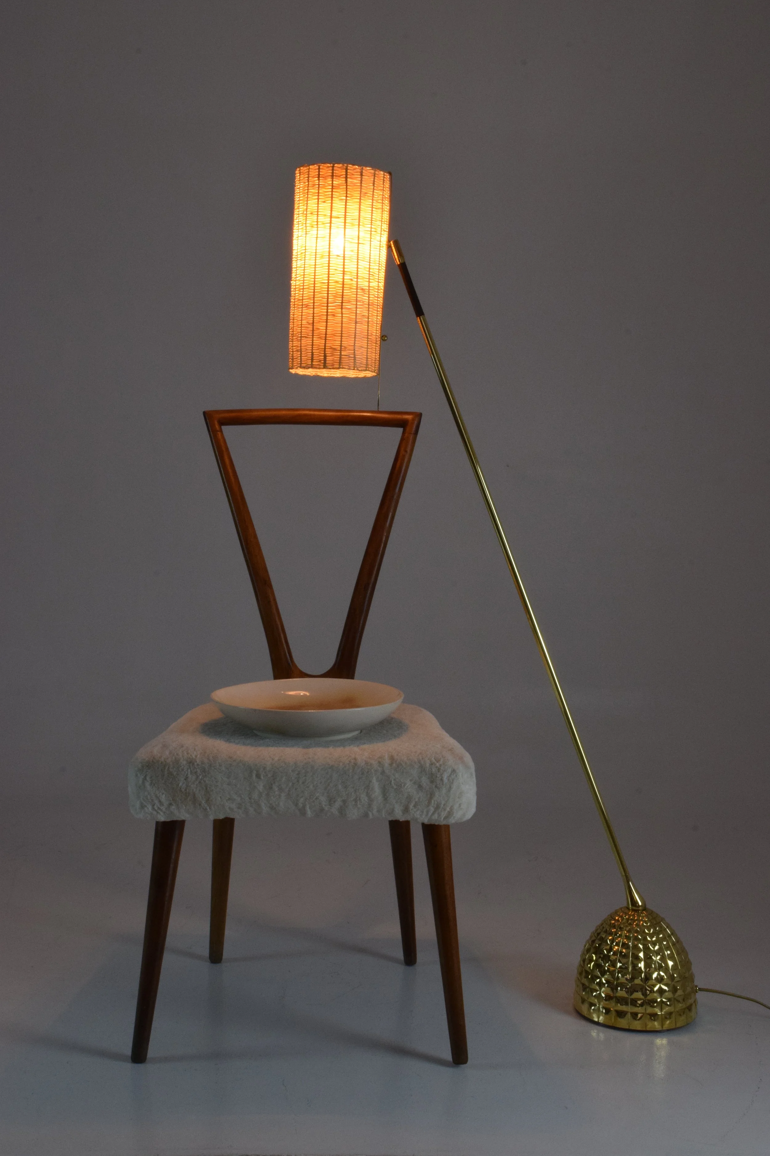 A modern chair with a wooden frame and a fluffy white seat cushion, a spherical white bowl on top, a tall lamp with a warm orange light, and a gold metallic floor lamp with a textured base, all arranged against a plain background.