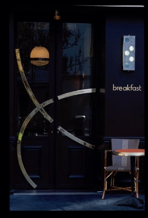 Dark entrance to a breakfast restaurant with wooden door and modern metal decorations, a chair and a small round table outside, and a sign with a light fixture reading "breakfast".


