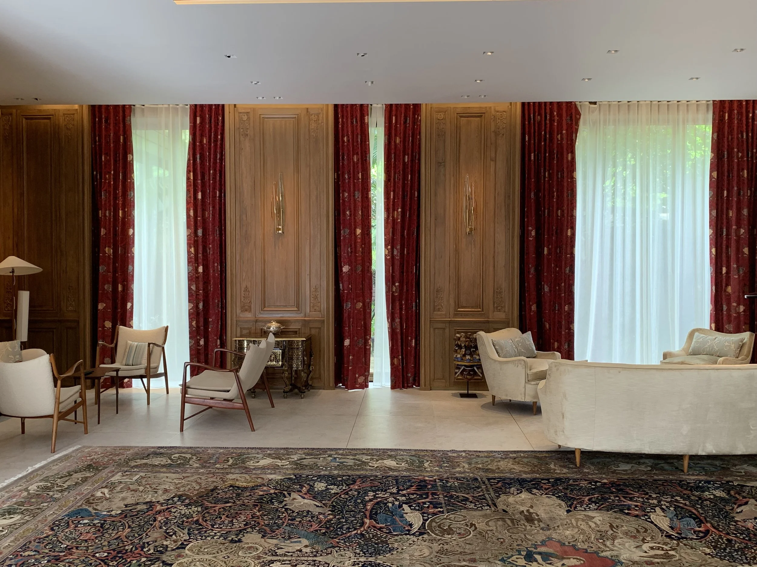 Elegant sitting area with cream-colored sofas and armchairs, red patterned curtains, and wooden wall panels, a Persian-style rug, and tall windows letting in natural light.


