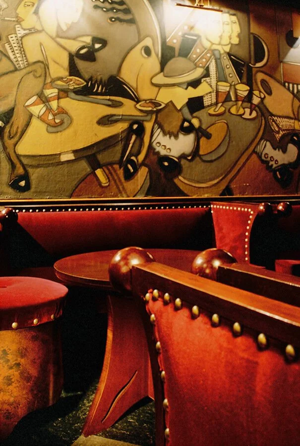 Dimly lit bar or restaurant with red upholstered furniture, including chairs and booths. A large, colorful mural with abstract human and animal figures decorates the wall above the seating area.


