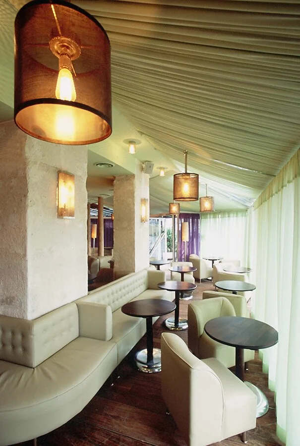 Interior of a restaurant or cafe with cream-colored seating, dark round tables, and multiple pendant lights hanging from a curved ceiling, with large curtains near the windows.