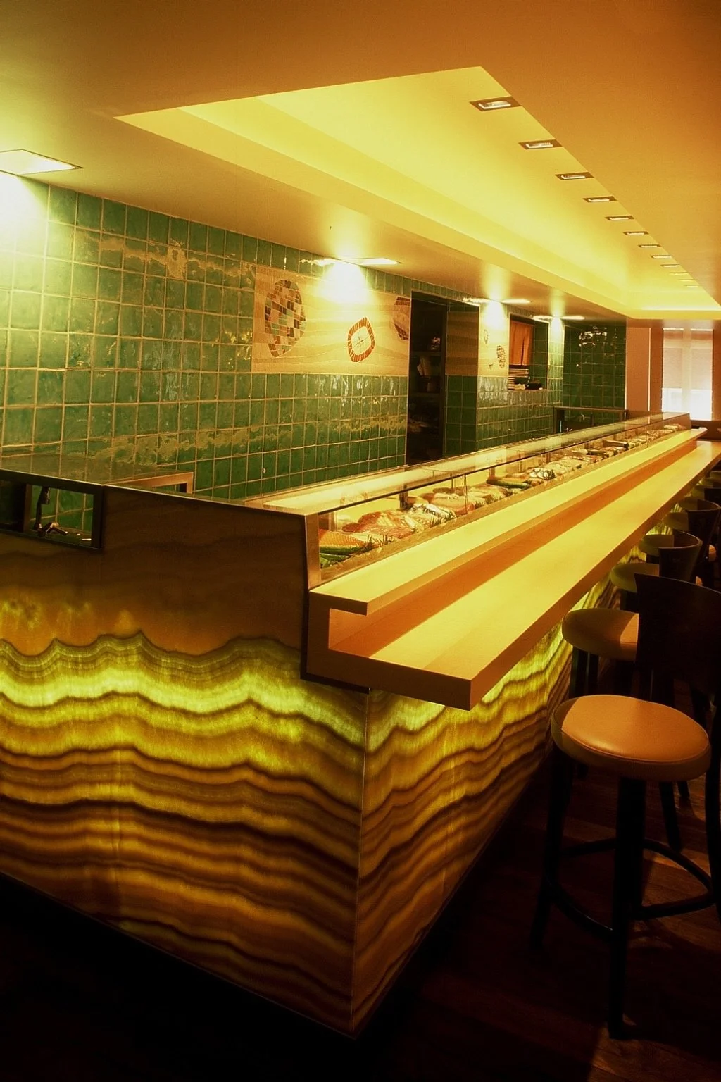 An illuminated bar with green tile walls, decorative mosaic art, and a backlit onyx or similar stone counter with bar stools.