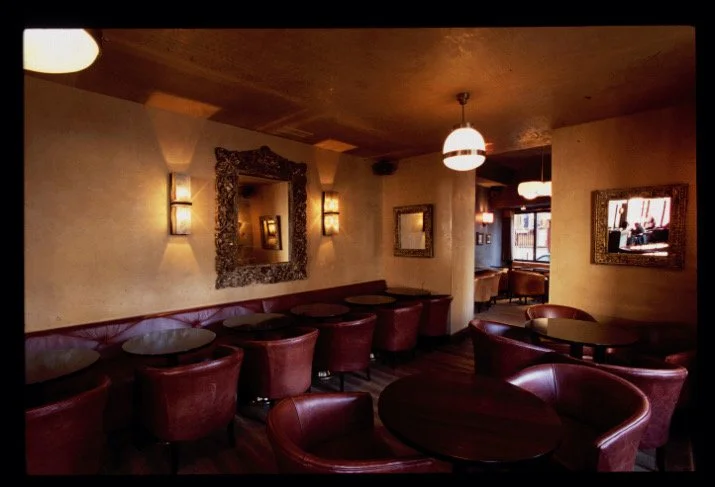 Interior of a cozy lounge with round tables, leather chairs, decorative mirrors on beige walls, and warm lighting.


