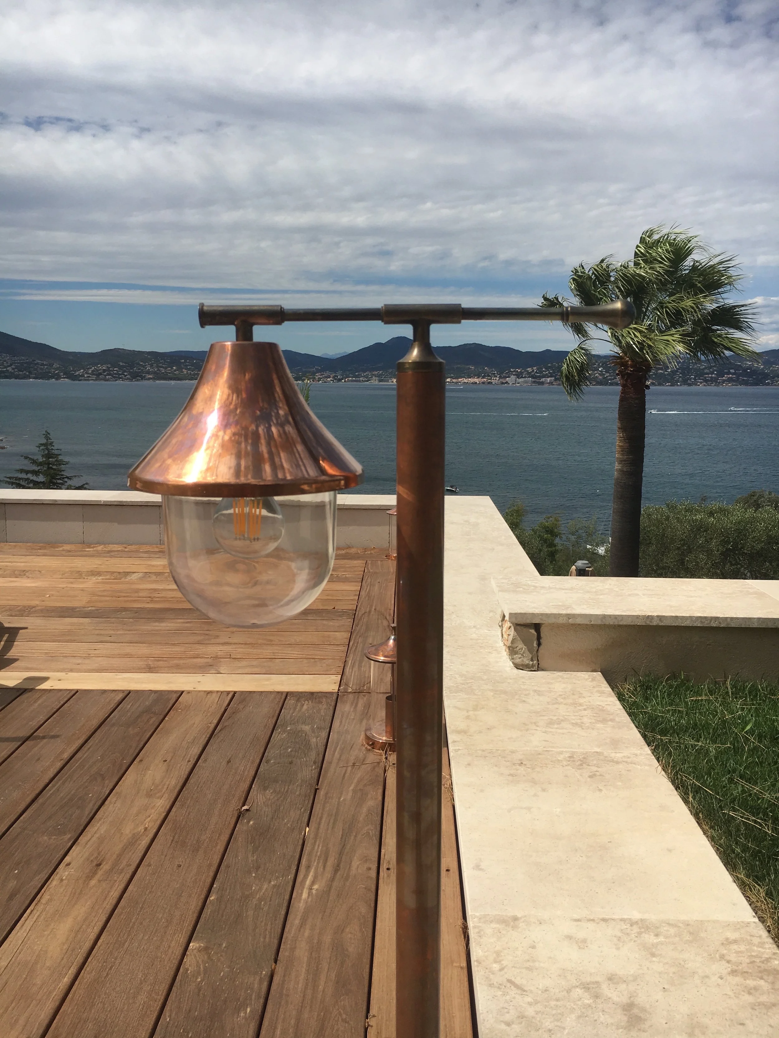 A copper and glass standing lamp with a bulb, outdoors on a wooden deck, with a scenic view of water, mountains, and a palm tree under a cloudy sky.


