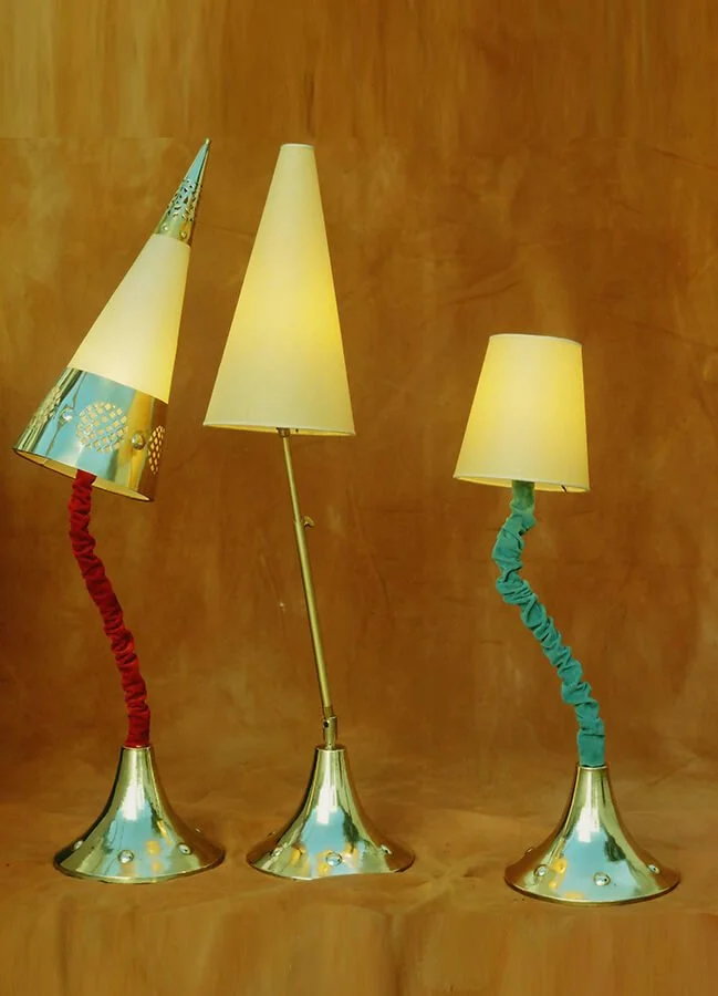 Three lamps with metallic bases and fabric-covered, curved, twisted lamp stems, each with a different shade, lined up against a wooden background.


