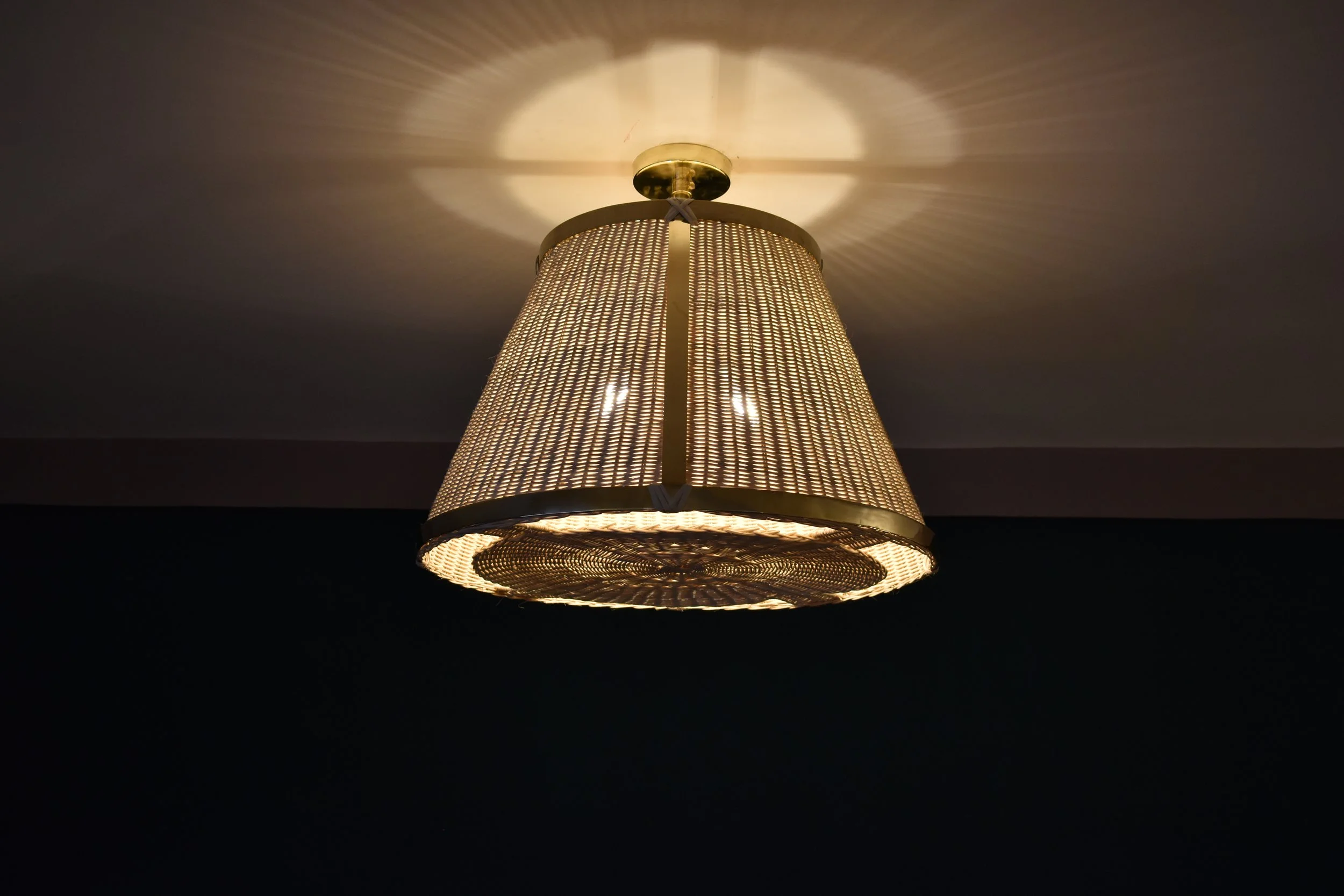 The Caeli-SD contemporary handcrafted made-to-measure pendant light fixture features an expertly handcrafted brass structure and a hand-woven dark-tinted rattan shade. 