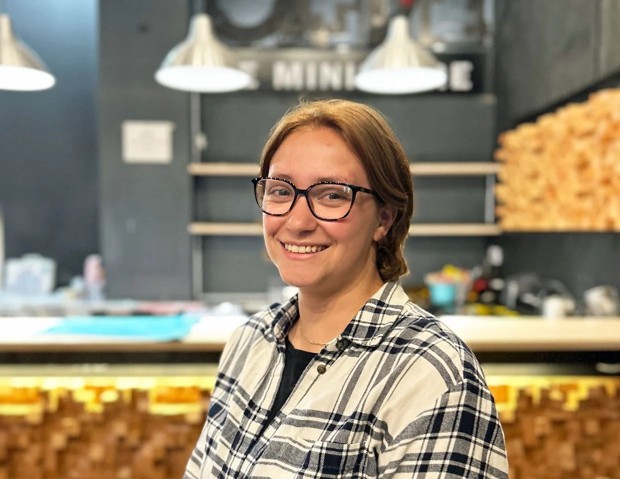 Une femme souriante portant des lunettes, habillée d'un cardigan à motifs, dans un intérieur moderne avec des lampes suspendues en arrière-plan.
