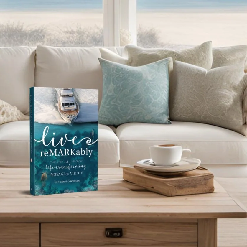 A white sofa with light-colored pillows in front of a window showing an outdoor view, a wooden coffee table with a book titled 'Live ReMARKably' leaning against a white cup and saucer on top of stacked wooden blocks.