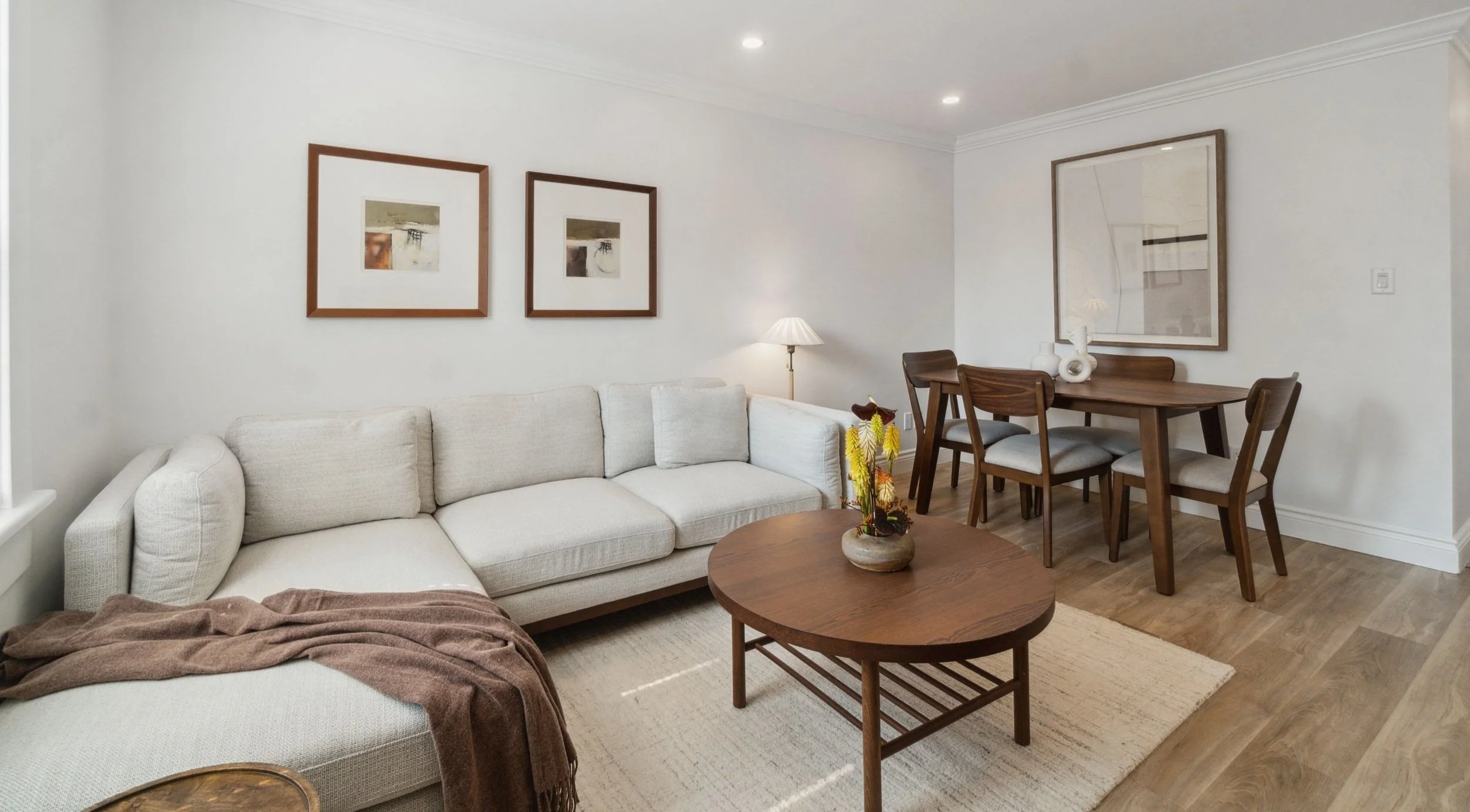 Living room with a white sectional sofa, a round wooden coffee table, framed abstract art on the walls, a floor lamp, and a wooden dining table with four chairs.
