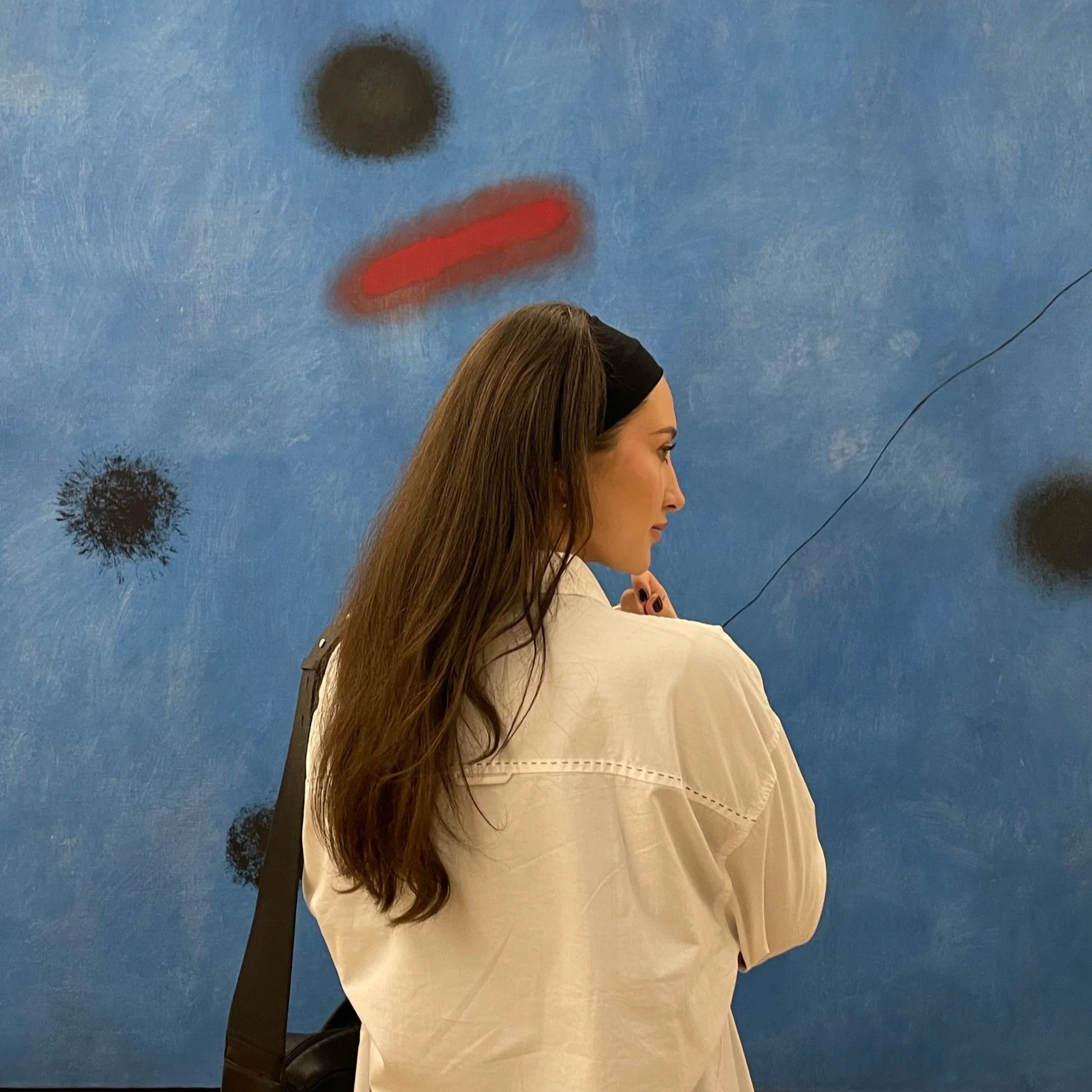 Side profile of a woman with long brown hair wearing a beige jacket and black headband, standing against a blue wall with abstract spray paint art of black and red shapes.