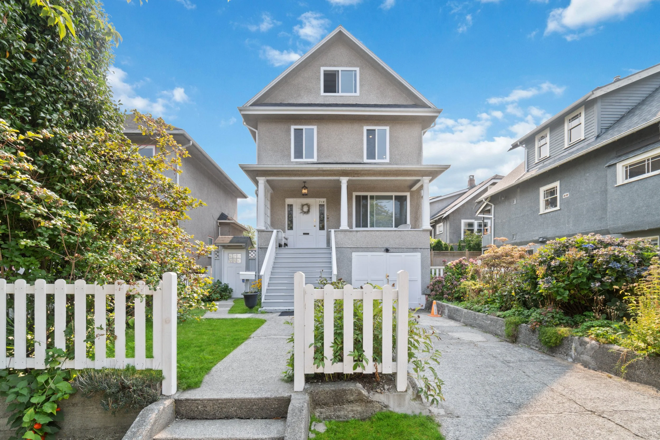 A white, three-story house with a front porch and a gabled roof, surrounded by a well-kept garden with a white picket fence, on a sunny day with a blue sky and scattered clouds.