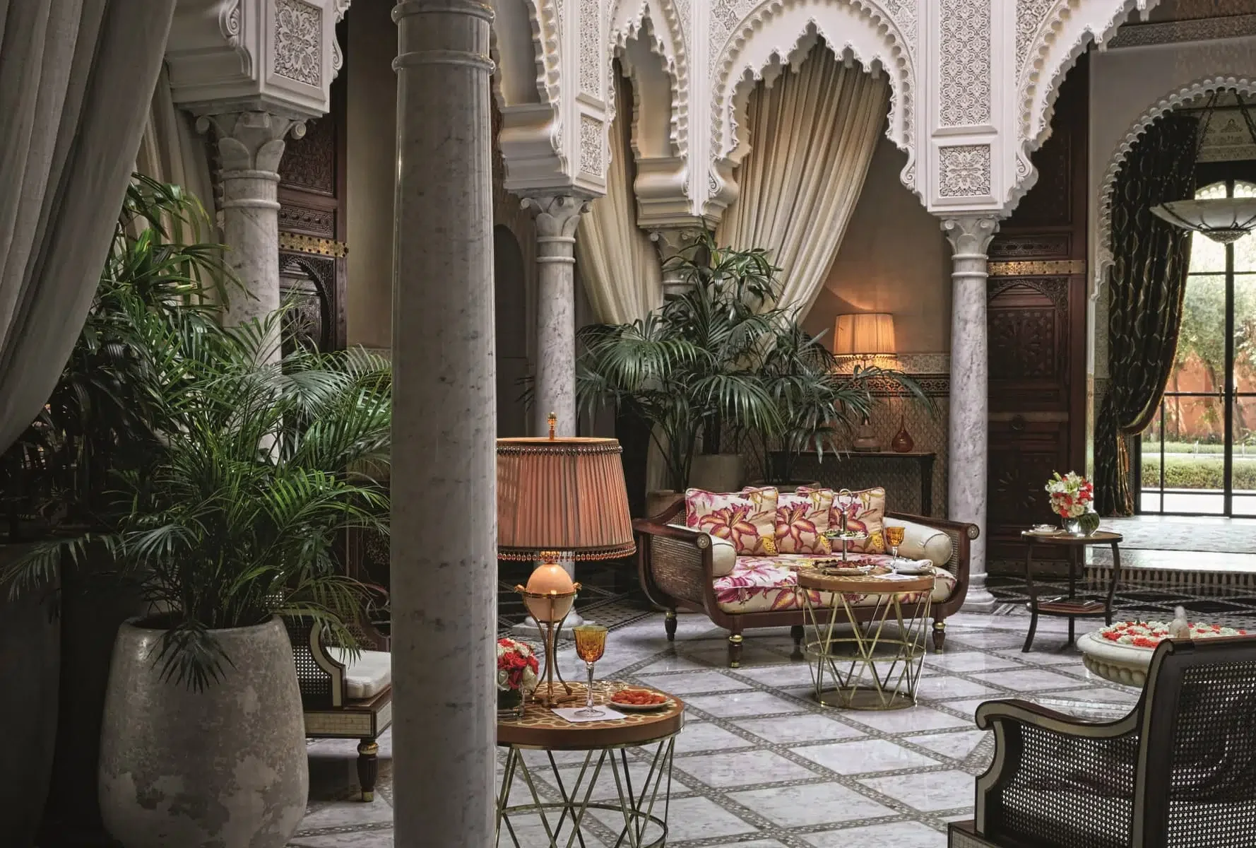 Elegant living room with marble columns, ornate arches, tropical plants, a floral sofa, side tables with lamps, and large windows letting in natural light.