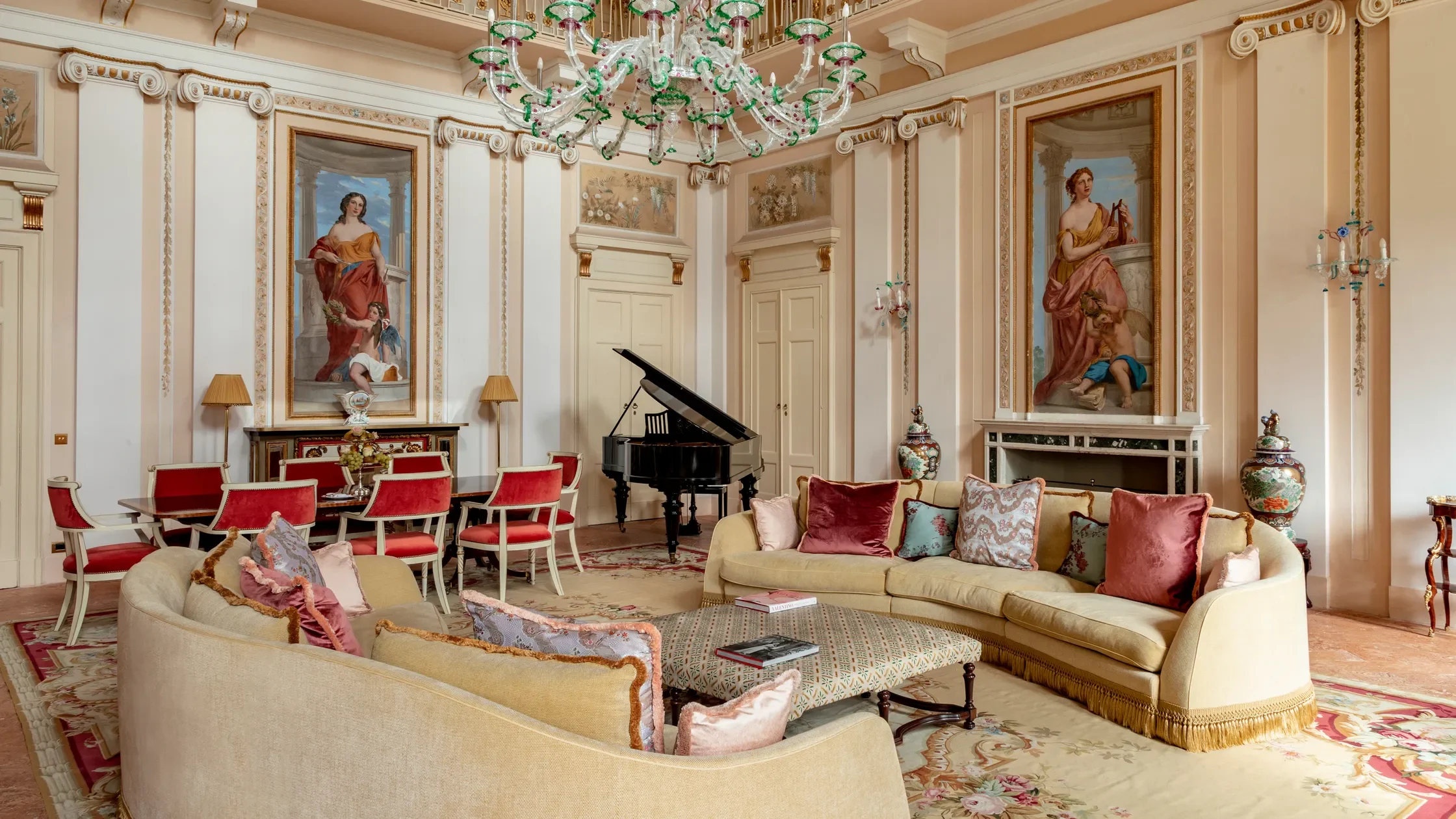 Luxurious living room with a grand chandelier, classical paintings, a black grand piano, a beige sofa with pink cushions, a long dining table with red and white chairs, and various decorative vases.