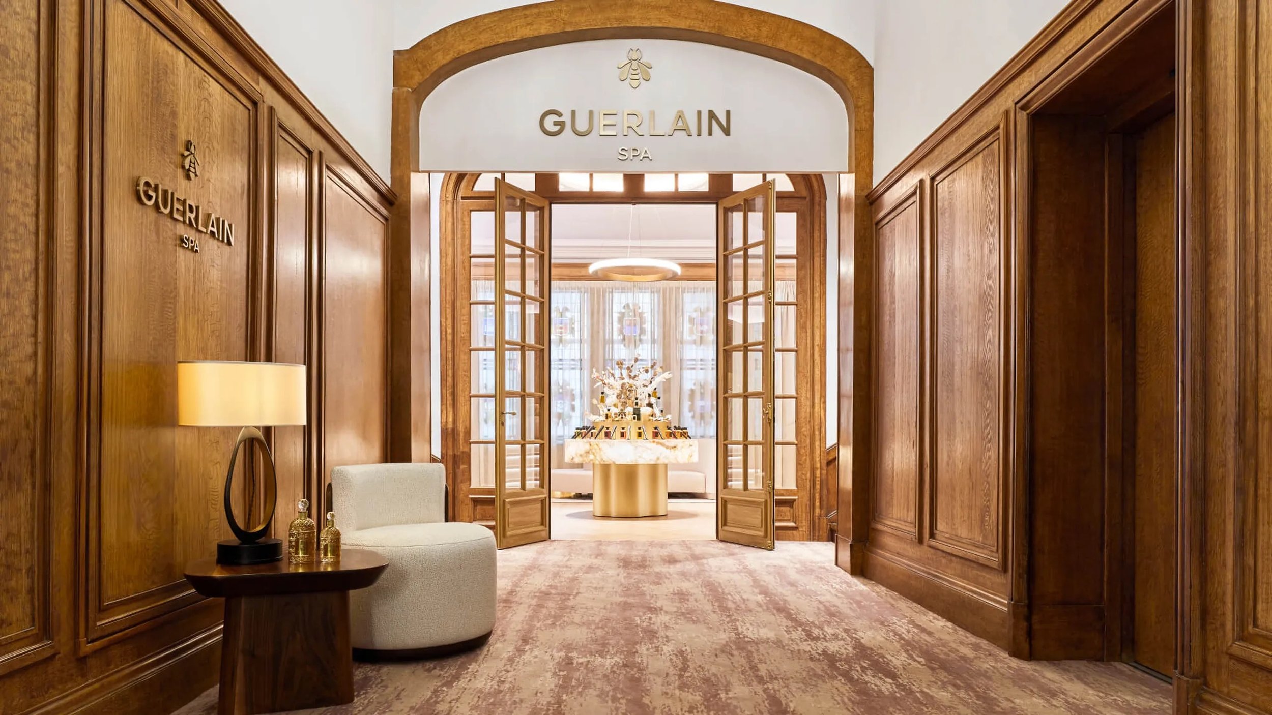 Wood-paneled entryway with open glass double doors leading to a room with a circular table, floral arrangement, and modern chandelier.