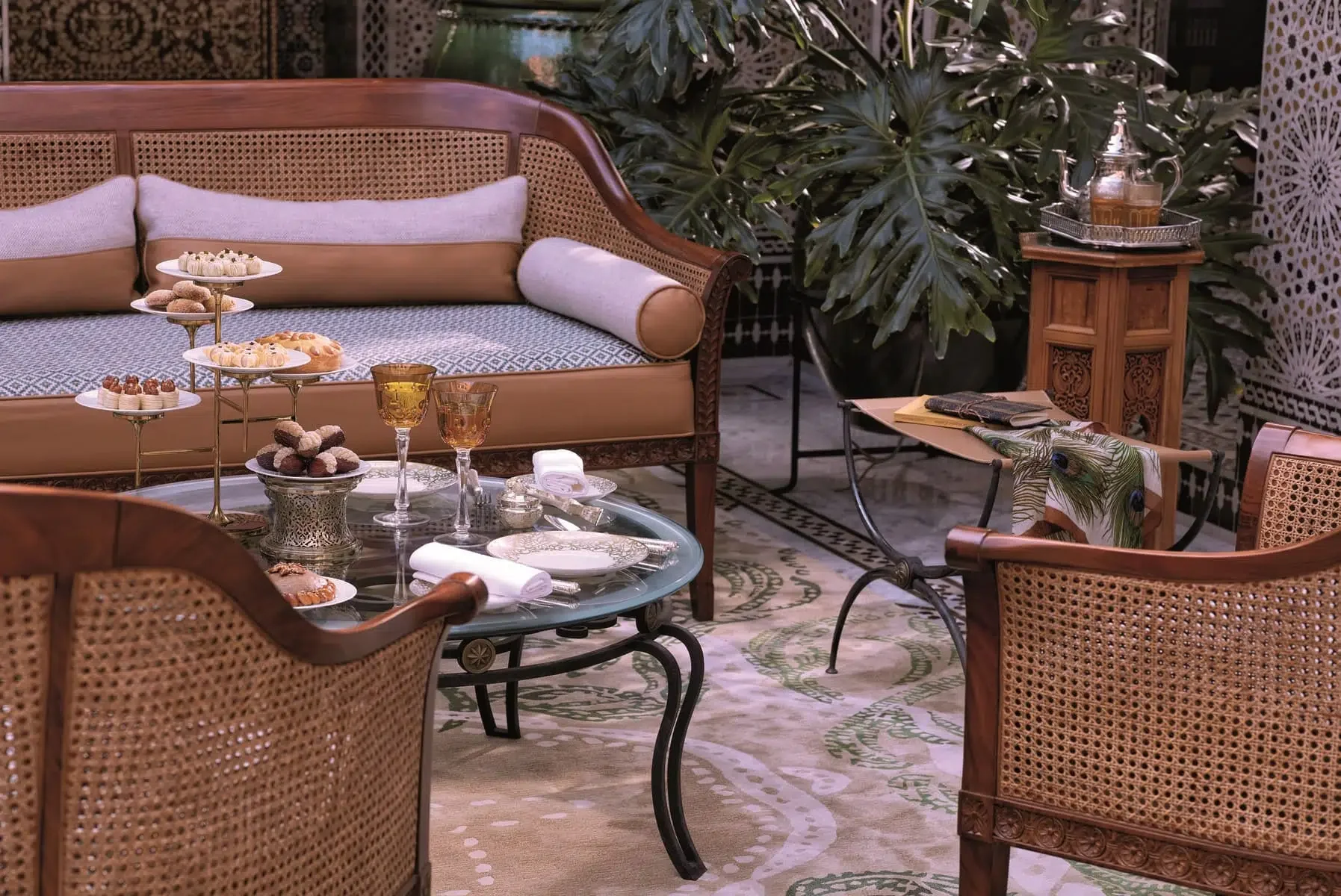 Elegant indoor setting with a wicker sofa, a glass coffee table with desserts and drinks, a wooden side table with a teapot, lush green plants, and decorative screens.