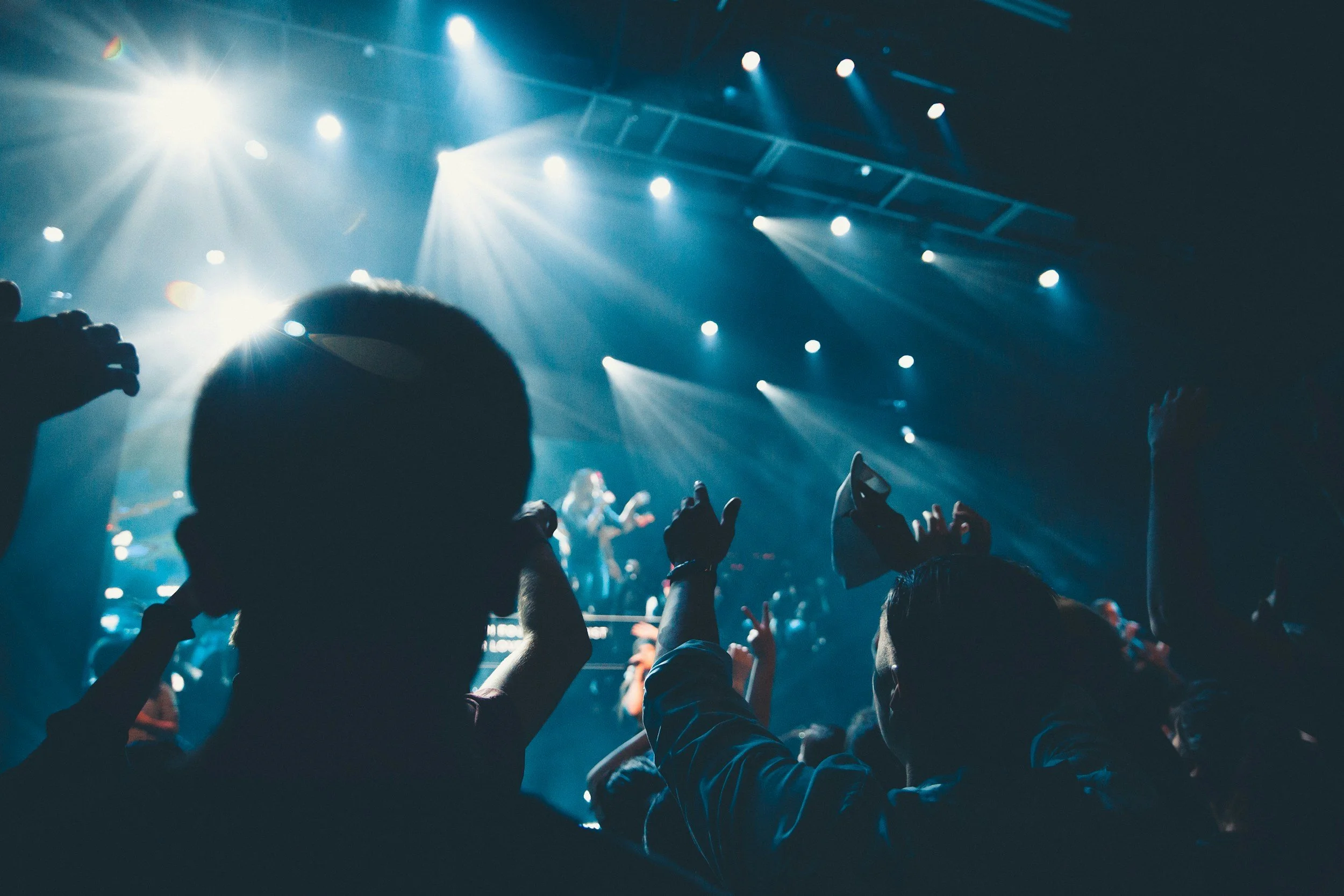 Concert scene with audience members raising their hands and taking photos, stage lighting, and performers in the background