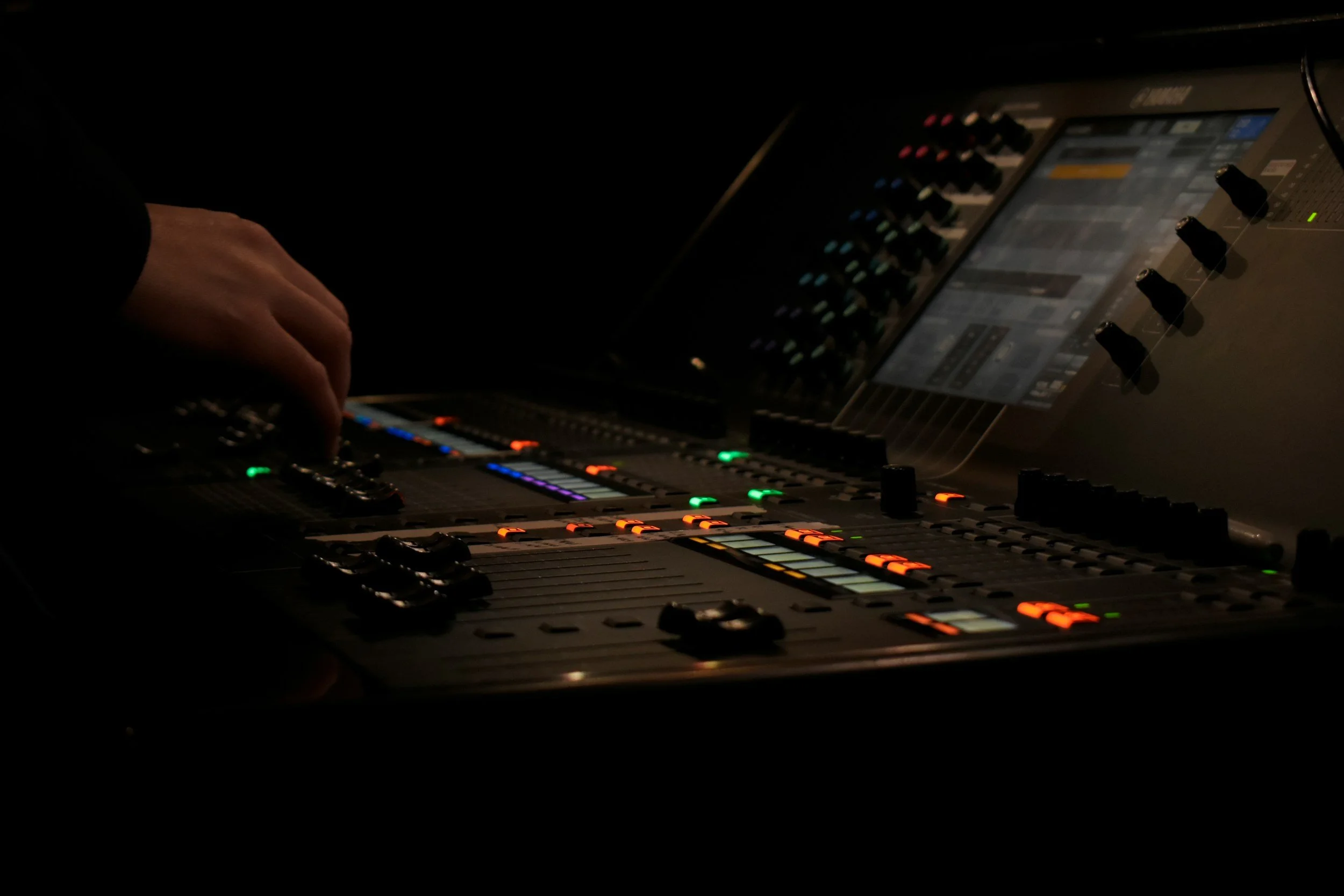 Close-up of a professional audio mixing console with illuminated buttons and faders, and a person's hand adjusting controls in a dark environment.