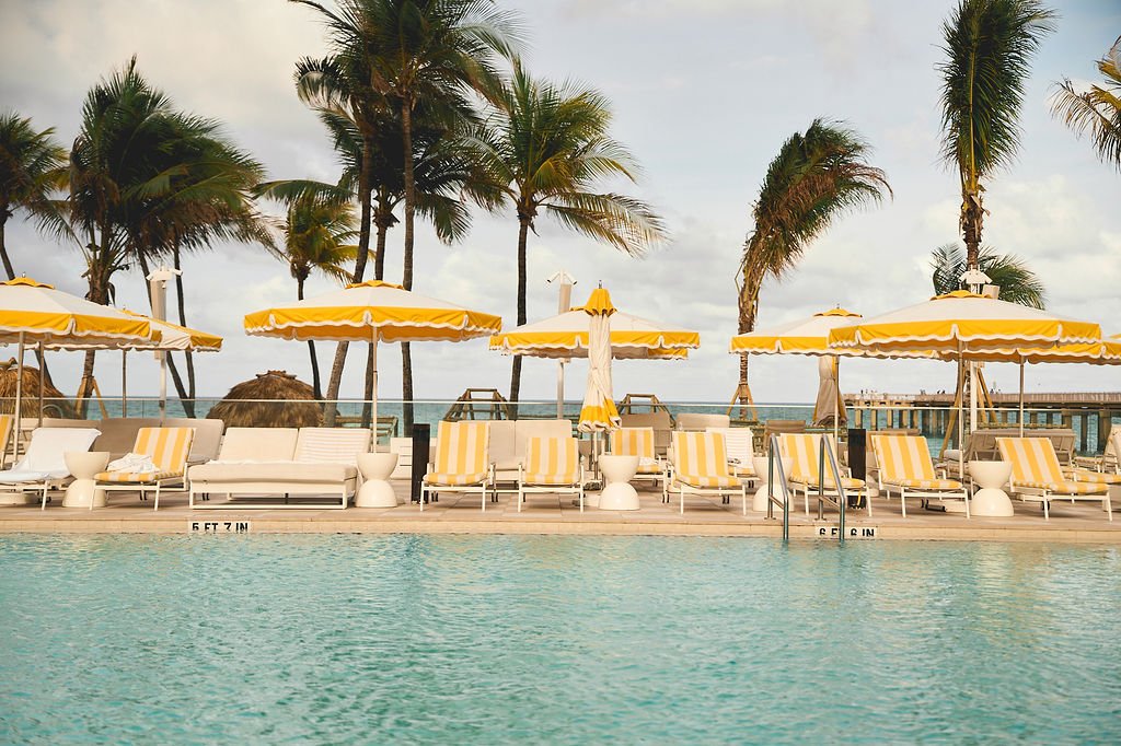 Swimming pool with yellow and white striped umbrellas and lounge chairs, palm trees, and a view of the ocean in the background