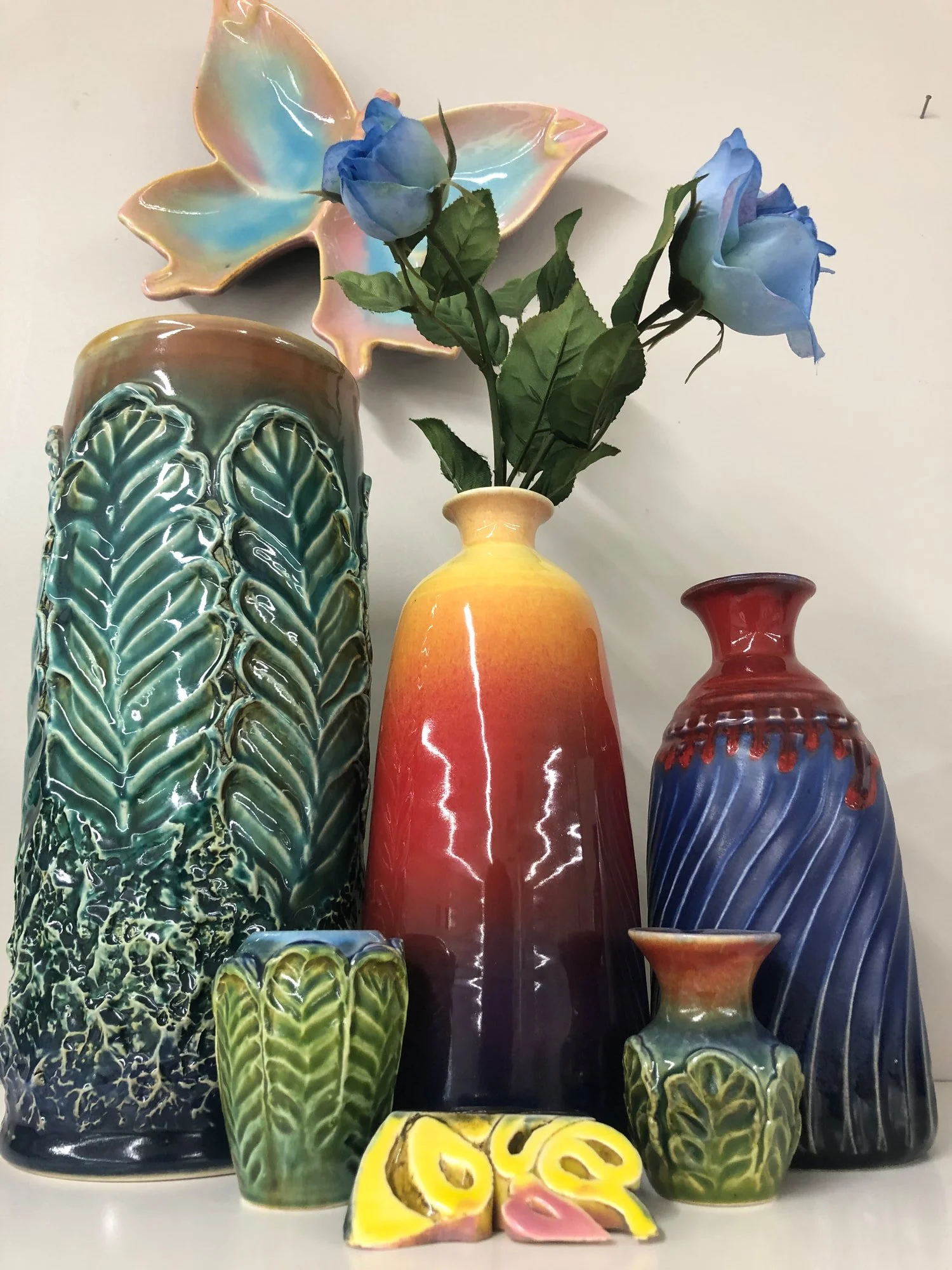Set of decorative ceramic vases, including a tall green and blue leaf-patterned vase, a yellow and red gradient vase with blue roses, a blue and red wavy vase, a small green leaf-patterned cup, a yellow and pink decorative piece, and a large butterfl