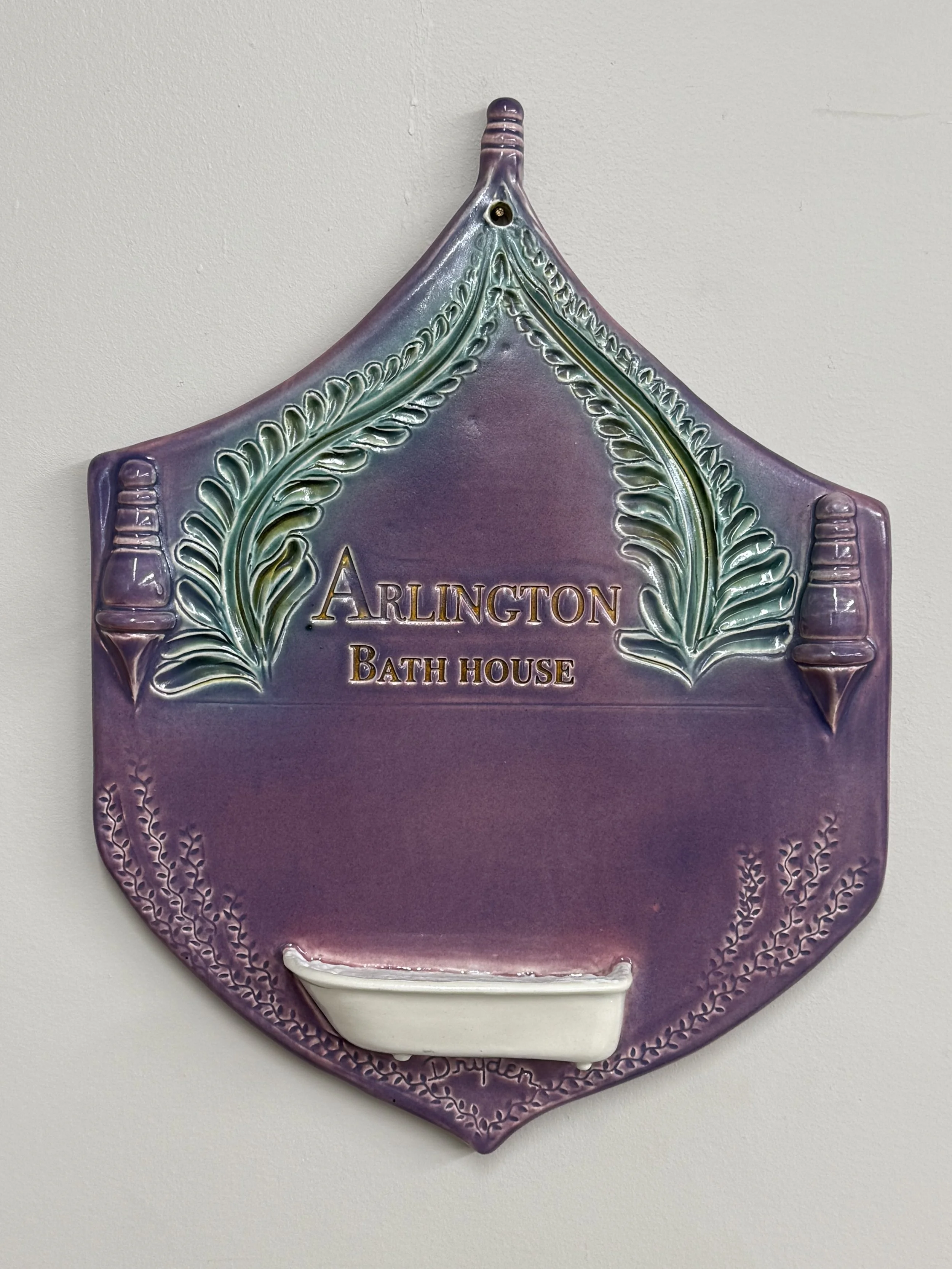 Decorative plaque for Arlington Bath House with a purple and green color scheme, leaf embellishments, and a small white soap dish at the bottom.