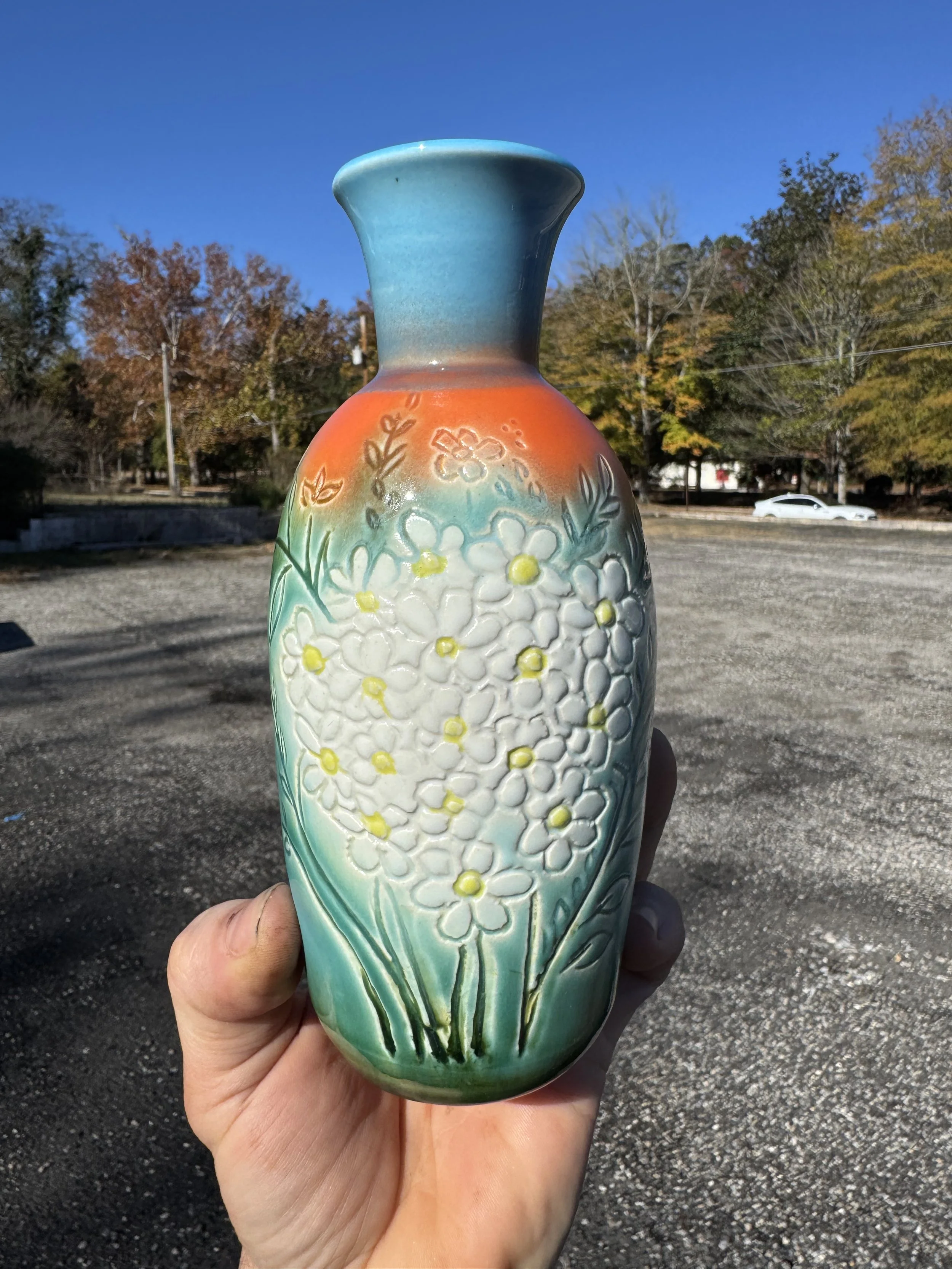 A person holding a ceramic vase with a floral embossed design, featuring white flowers with yellow centers, against a background of trees and a clear blue sky.