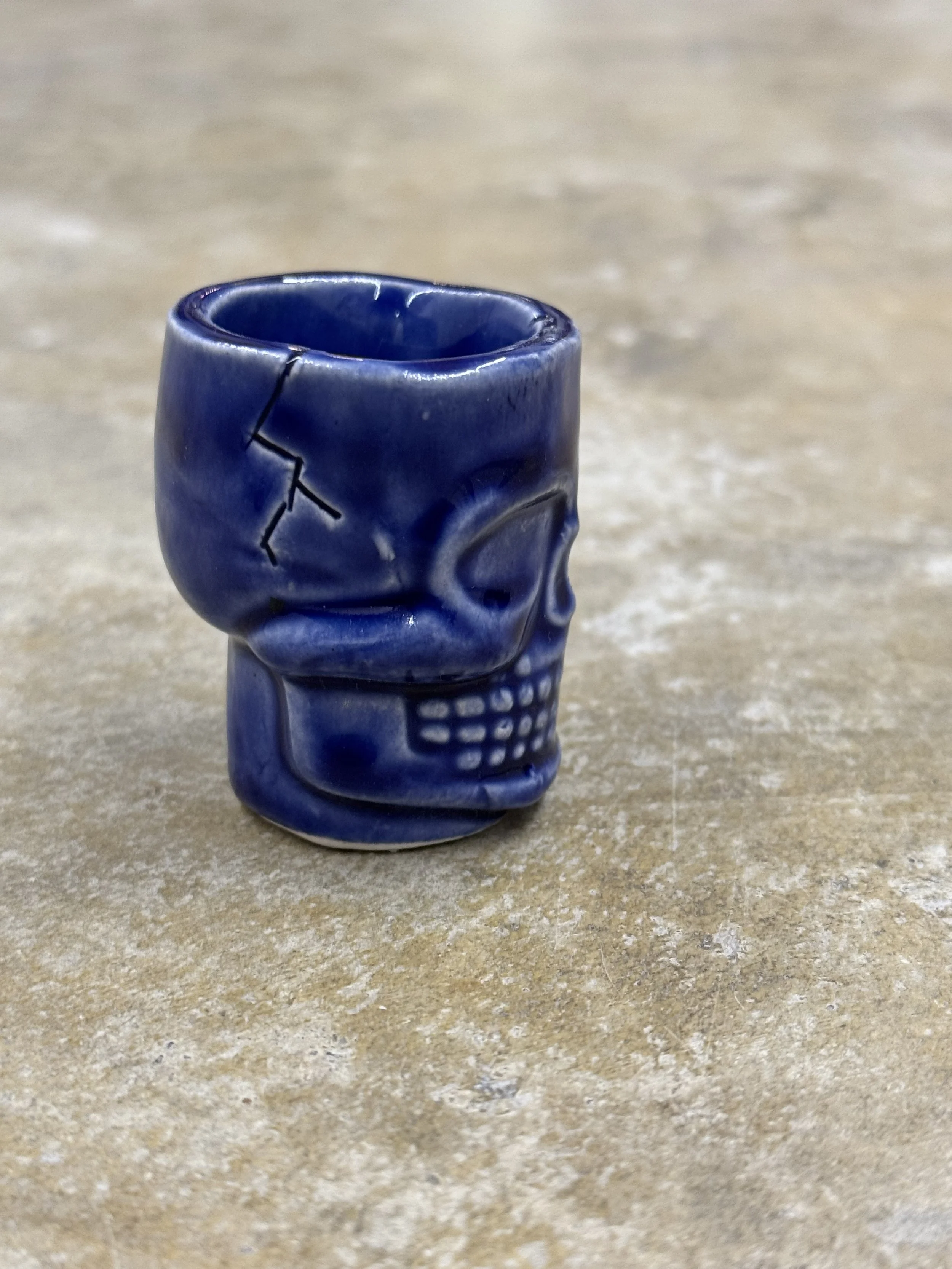 A small blue skull-shaped cup with a cracked design, placed on a beige textured surface.