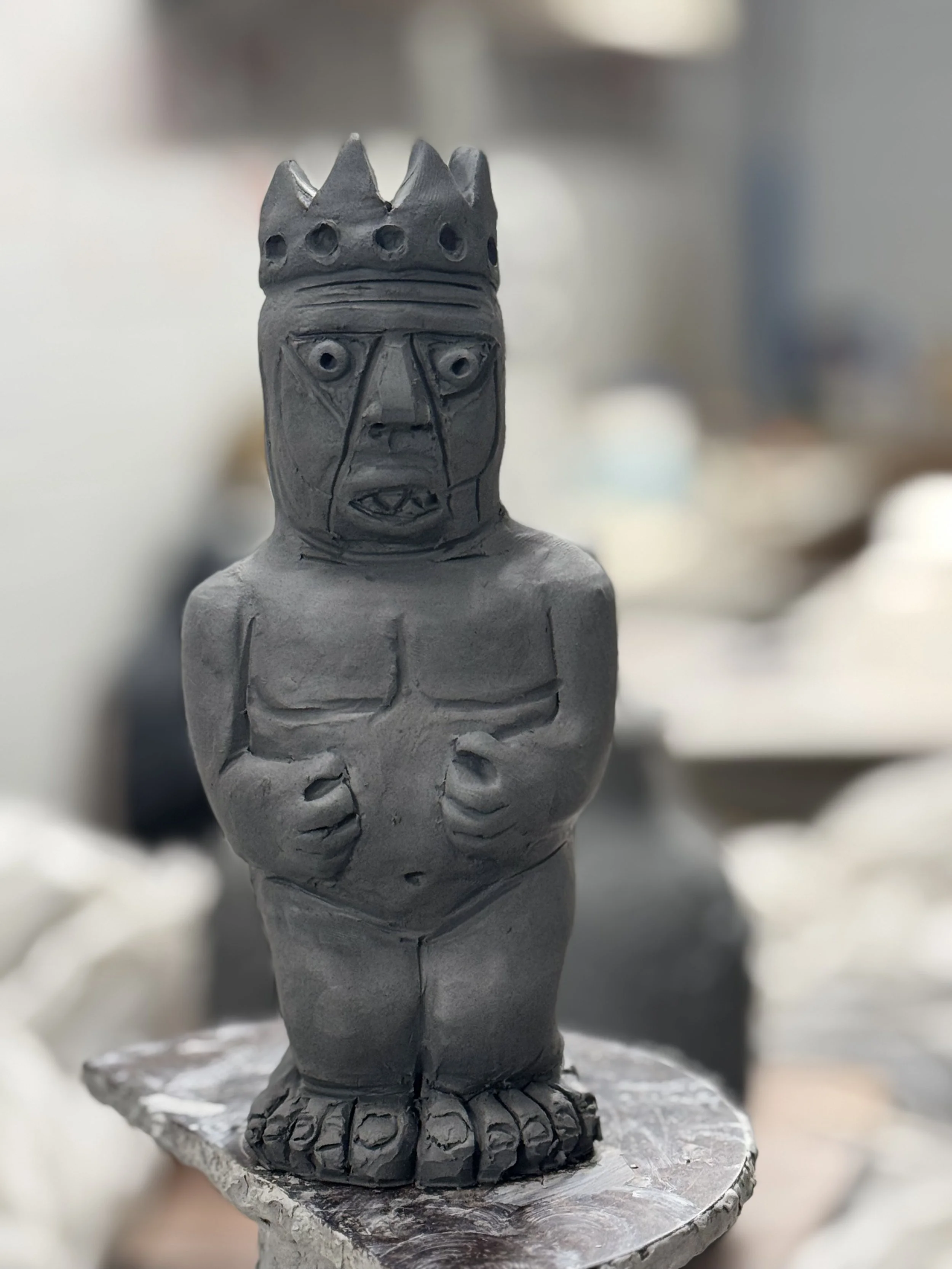 A gray clay sculpture of a humanoid figure with a crown, exaggerated facial features, and clenched fists, standing on a circular base.