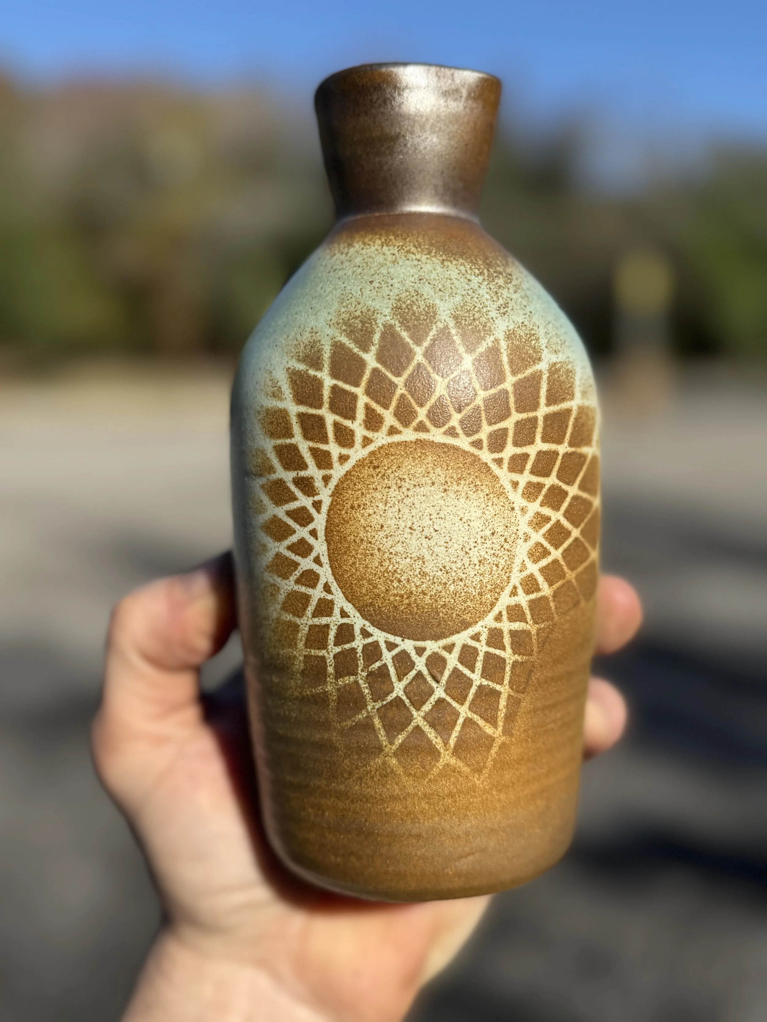 A person holding a ceramic vase with a geometric pattern and a sun design in earthy tones, with a blurred outdoor background.