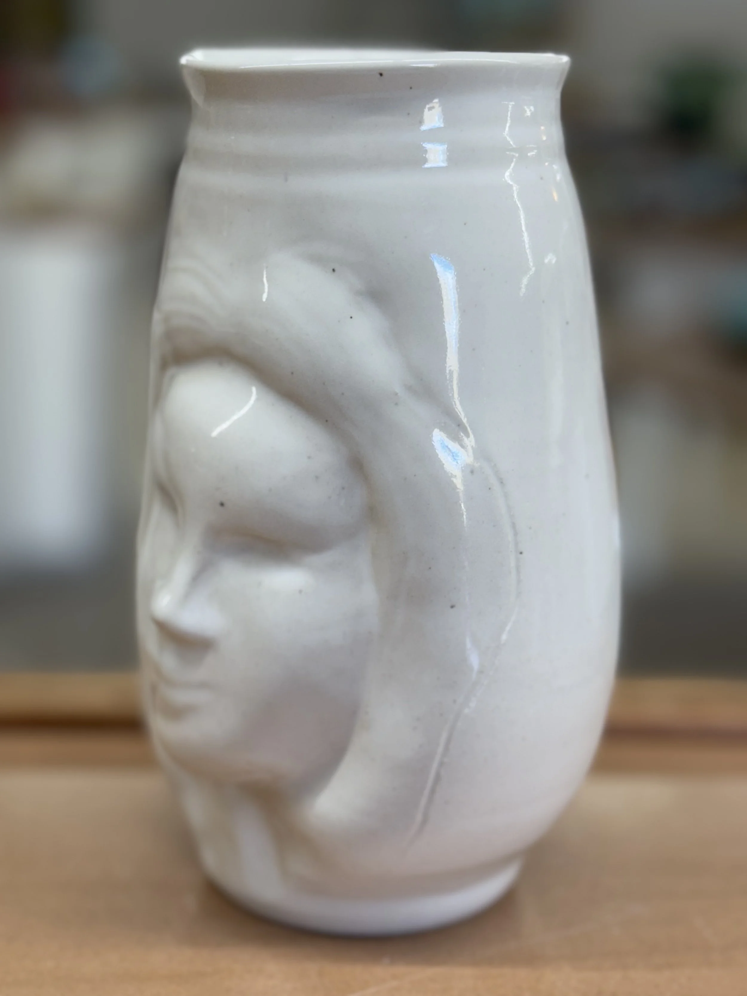 White ceramic vase with a raised facial profile, featuring closed eyes and full lips, placed on a wooden surface.