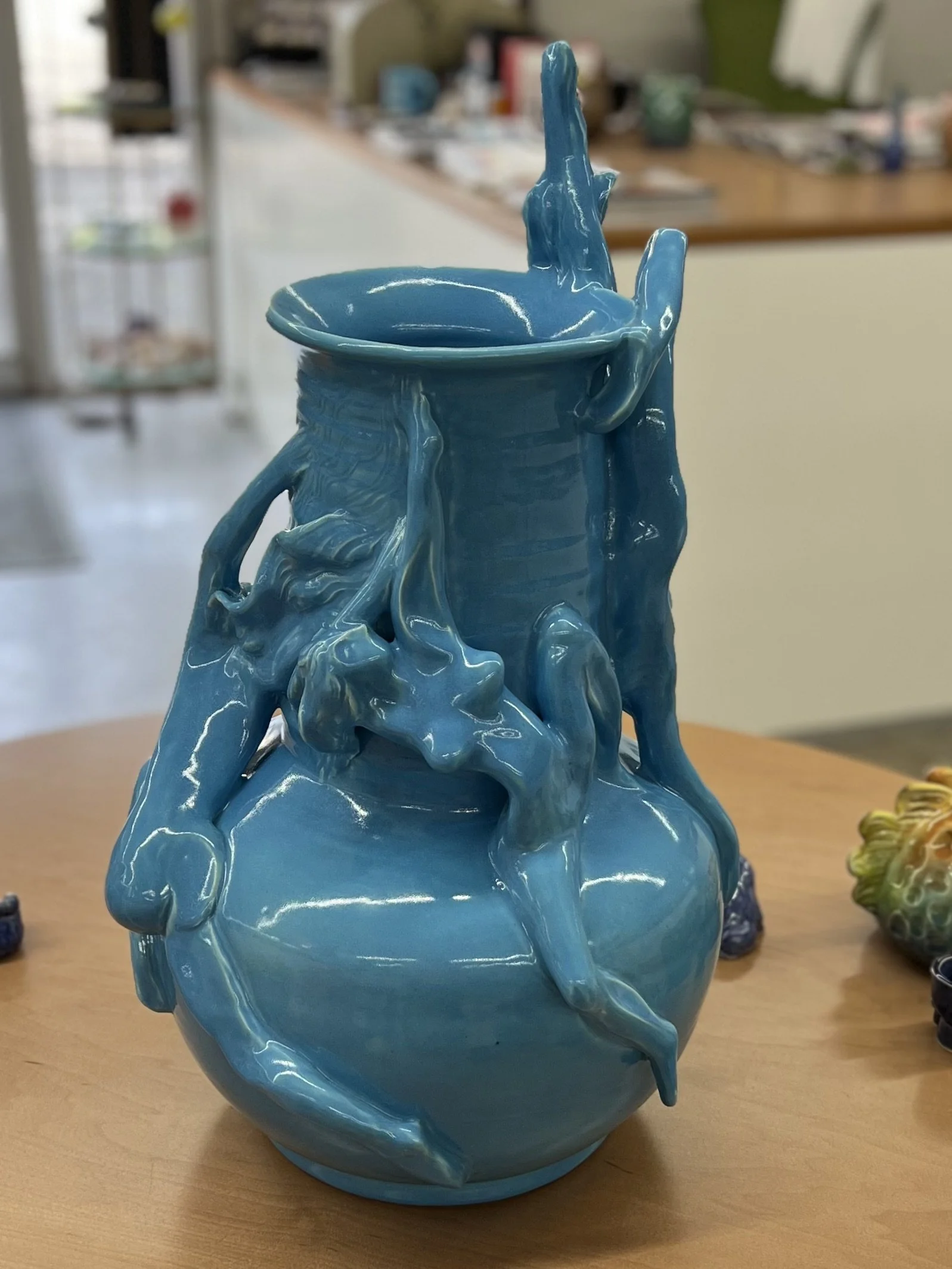 A glossy blue ceramic vase with intricate, sculpted female figures that resemble flowing liquid or twisting vines, placed on a wooden surface.