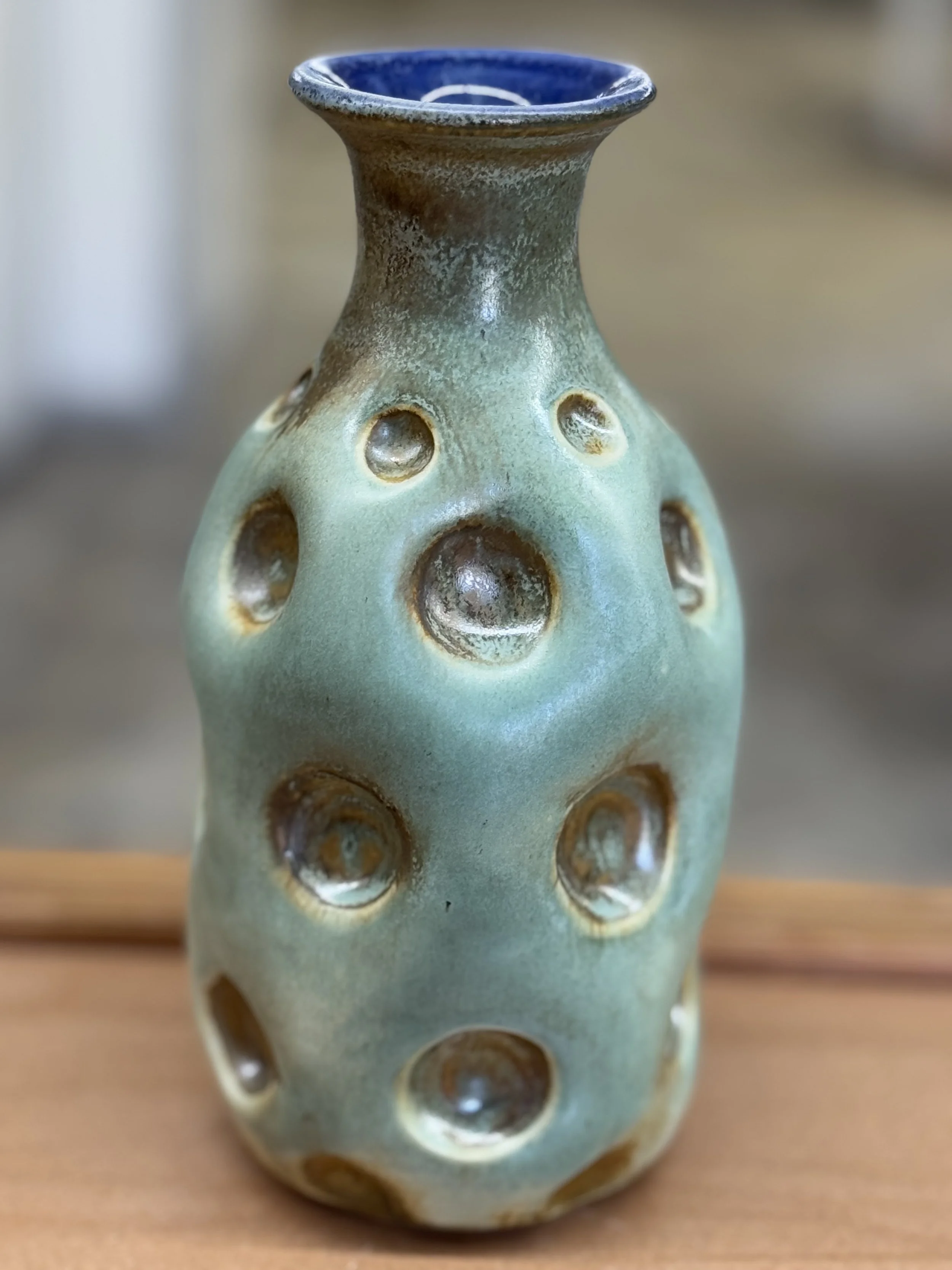 Ceramic vase with multiple round holes, glazed with green and blue tones, placed on a wooden surface.