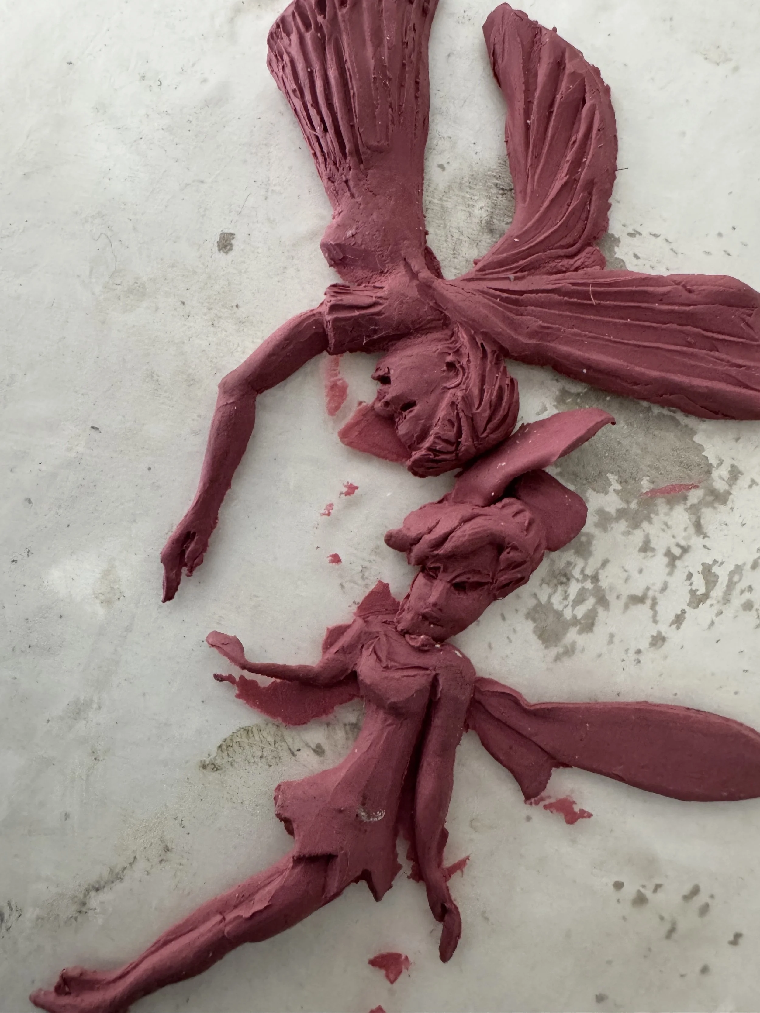 A pinkish-red sculpture of a fairy with wings sitting on a flat surface.