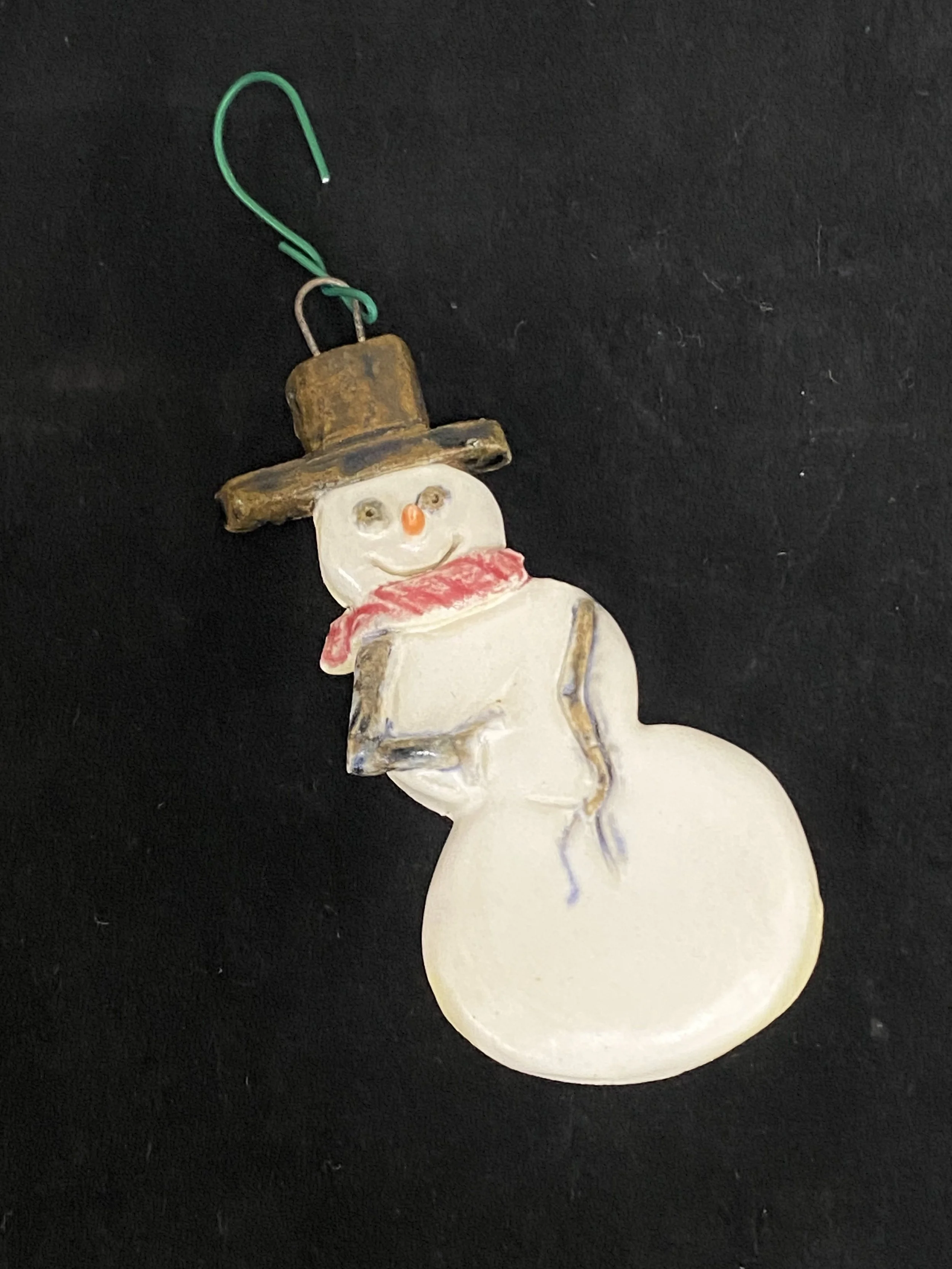 A snowman ornament with a rusted top hat, red scarf, and a small book, attached by a green wire hook.