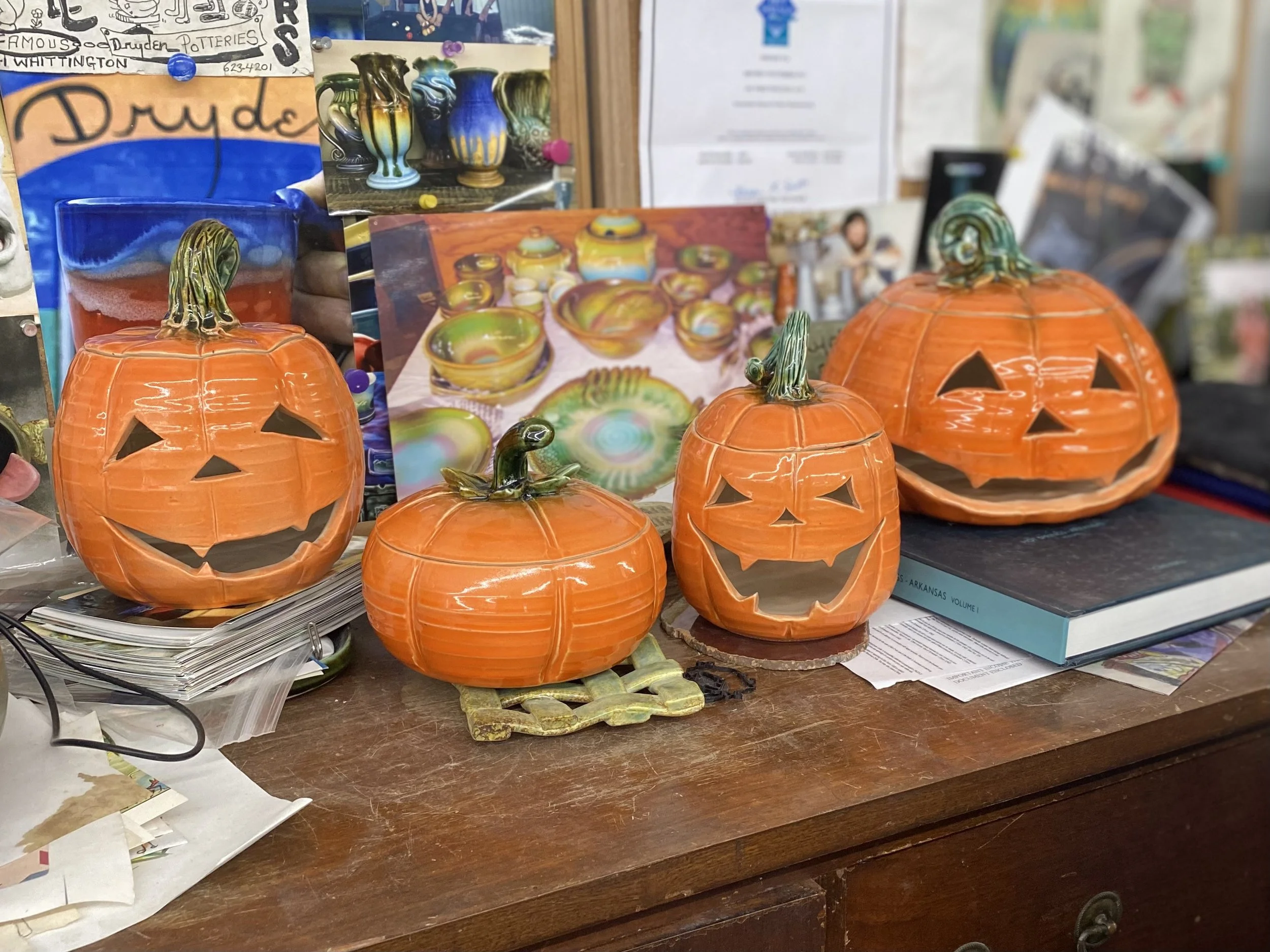 Four orange ceramic pumpkin lanterns with carved faces, sitting on a cluttered wooden desk among papers, books, photographs, and colorful pottery images in the background.
