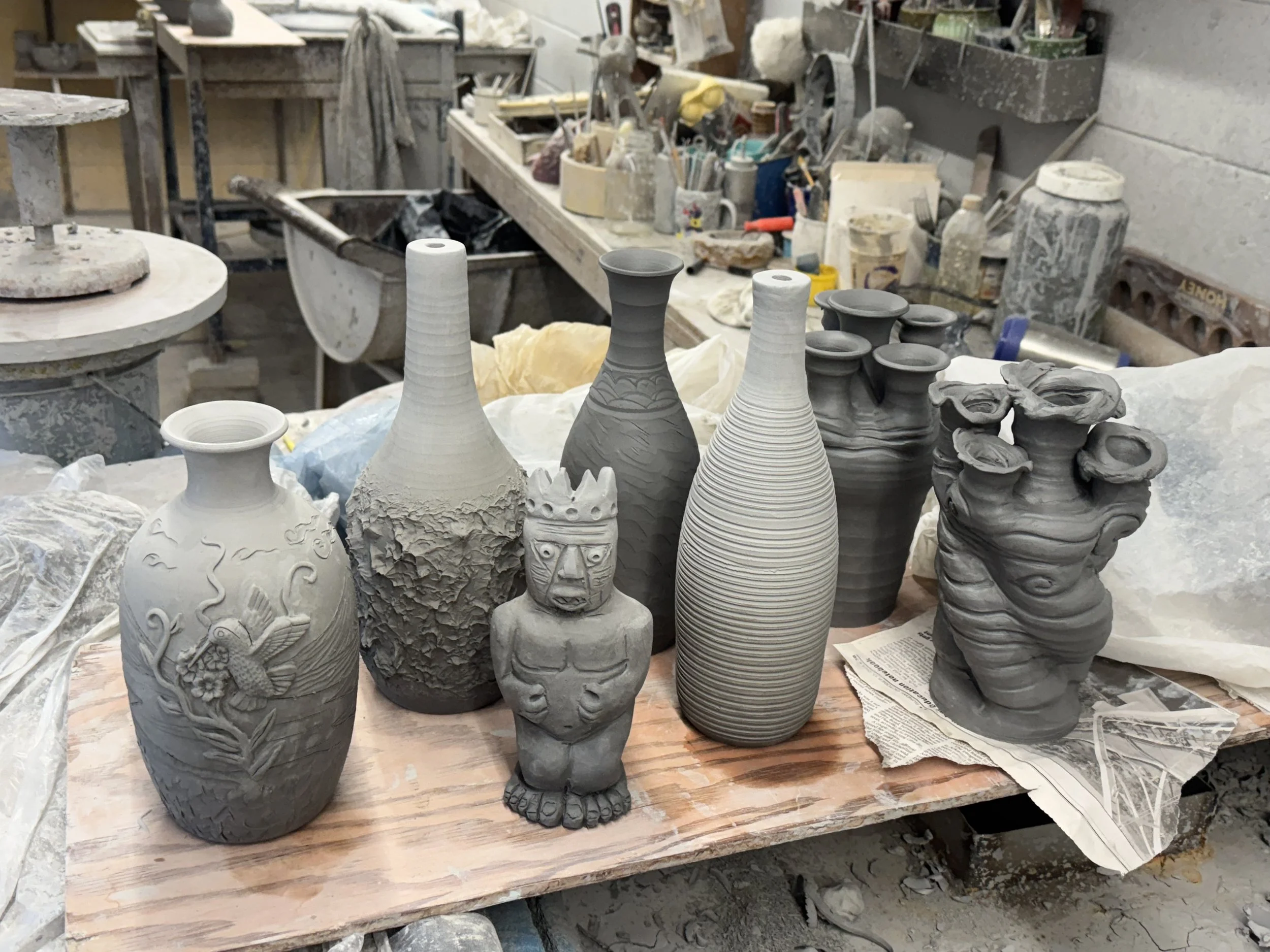 A collection of unfinished ceramic vases and a figurine with a tribal design, placed on a wooden surface in a pottery workshop.