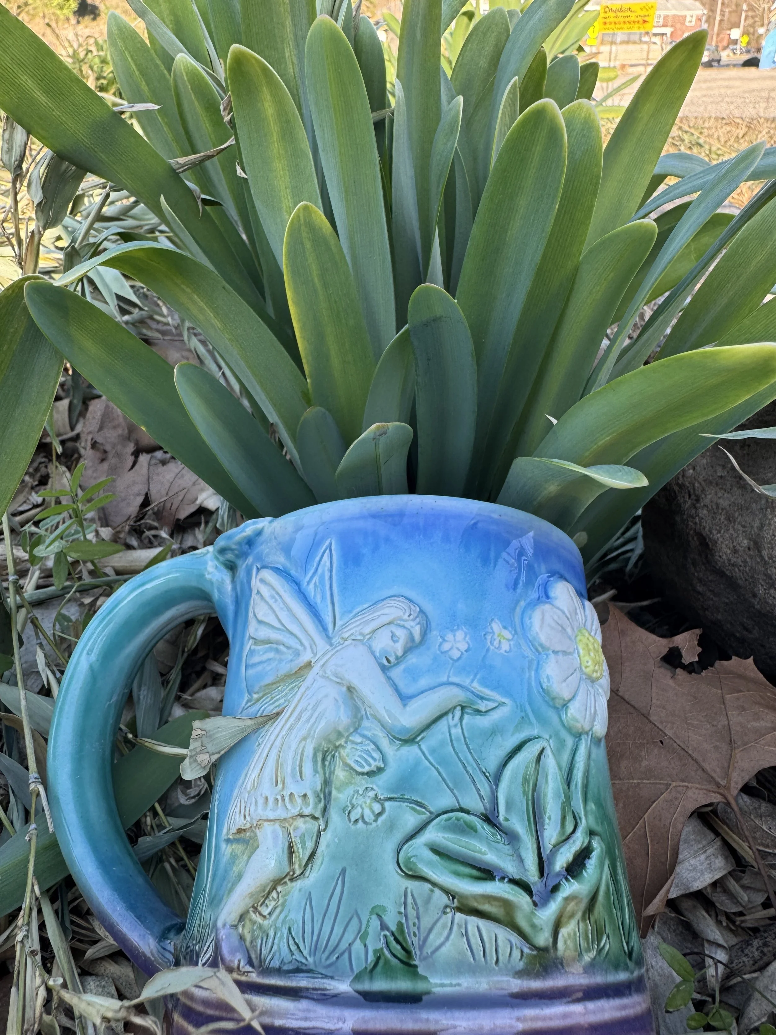 Close-up of a decorative ceramic mug with a fairy design, placed in front of a large green plant with long, pointed leaves, outdoors.