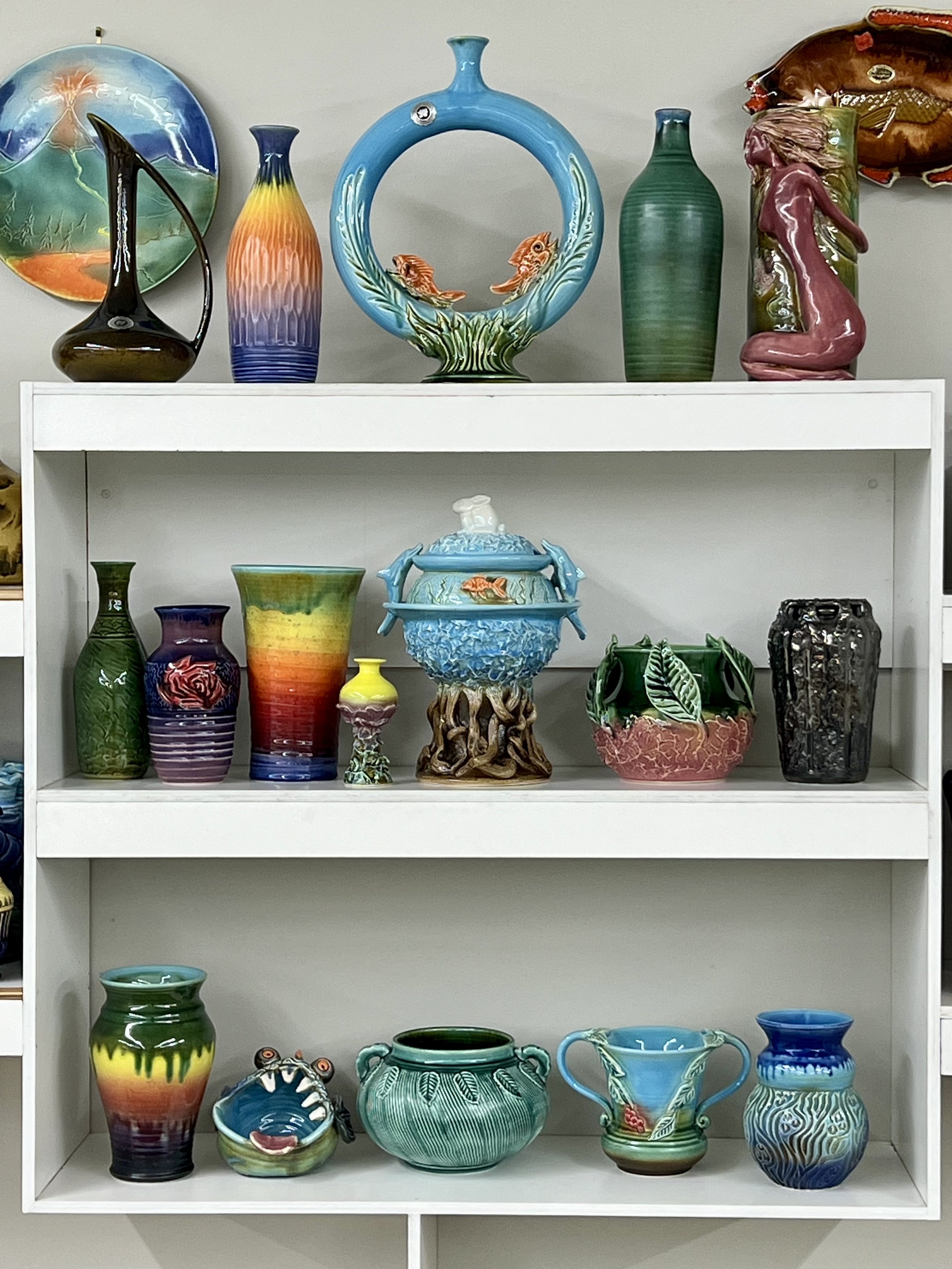 Colorful decorative vases and ceramic sculptures displayed on a white shelf, with various shapes and designs including fish, flowers, abstract figures, and a dragon.