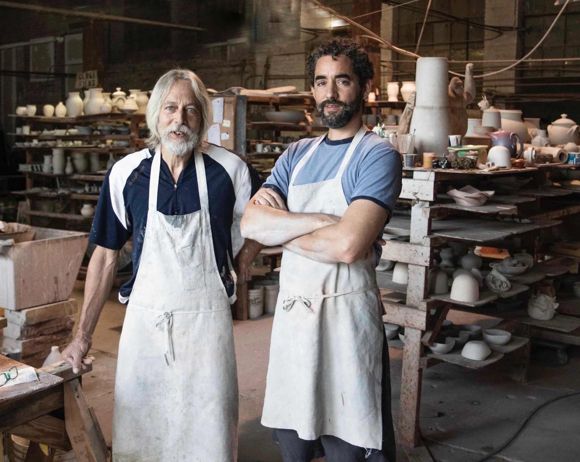 Two men standing inside a ceramics studio, wearing aprons, with shelves of pottery and clay objects in the background.