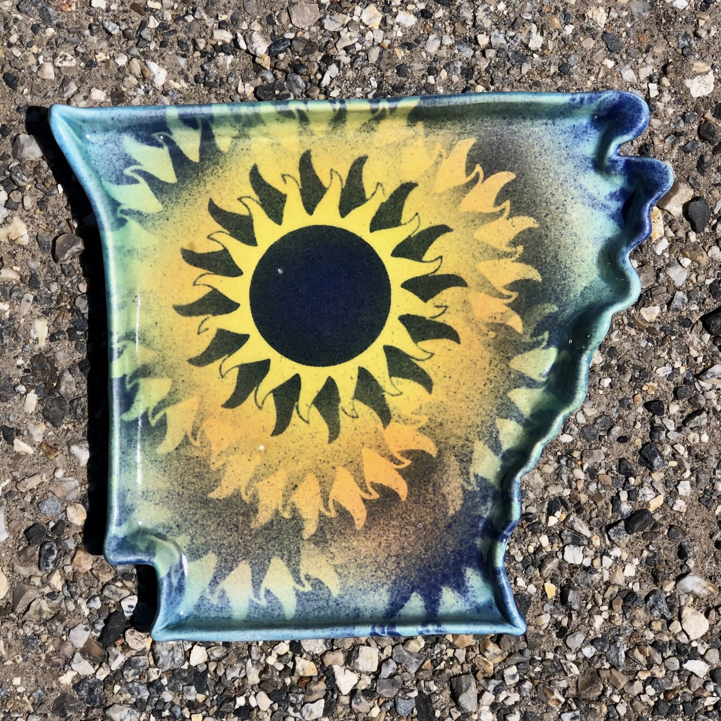 Decorative ceramic ashtray with a black sun design on a green and yellow background, placed on gravel ground.