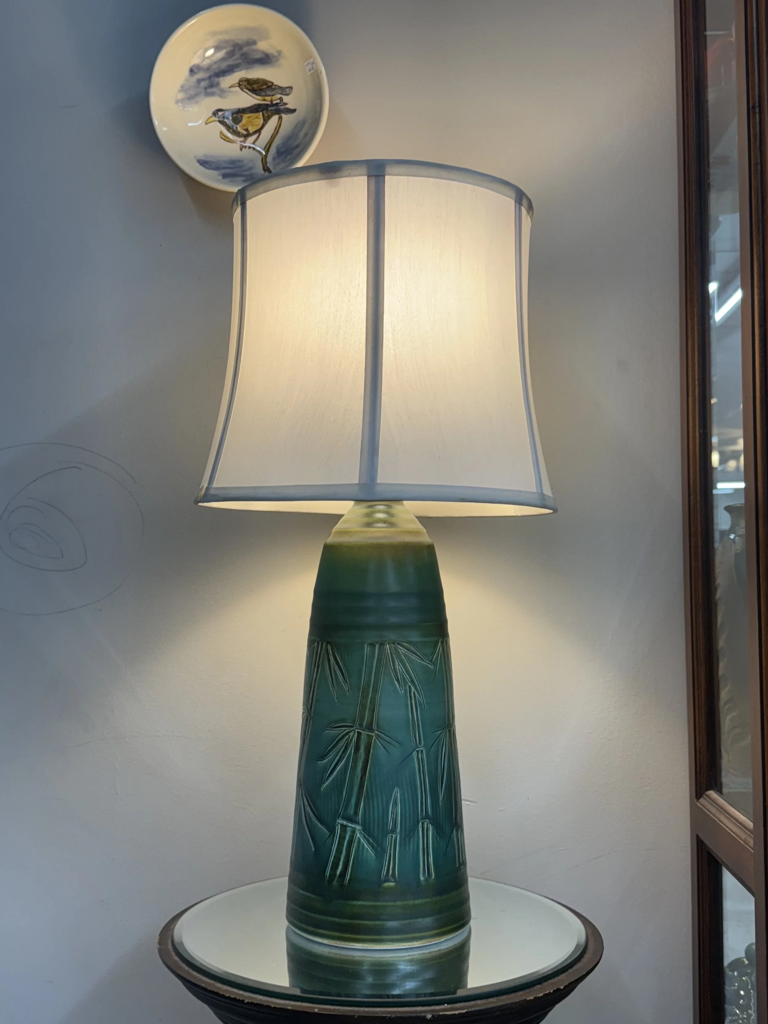 A ceramic table lamp with a green bamboo design, topped with a white fabric lampshade, placed on a round glass table. A decorative plate with birds is hung on the wall behind the lamp.