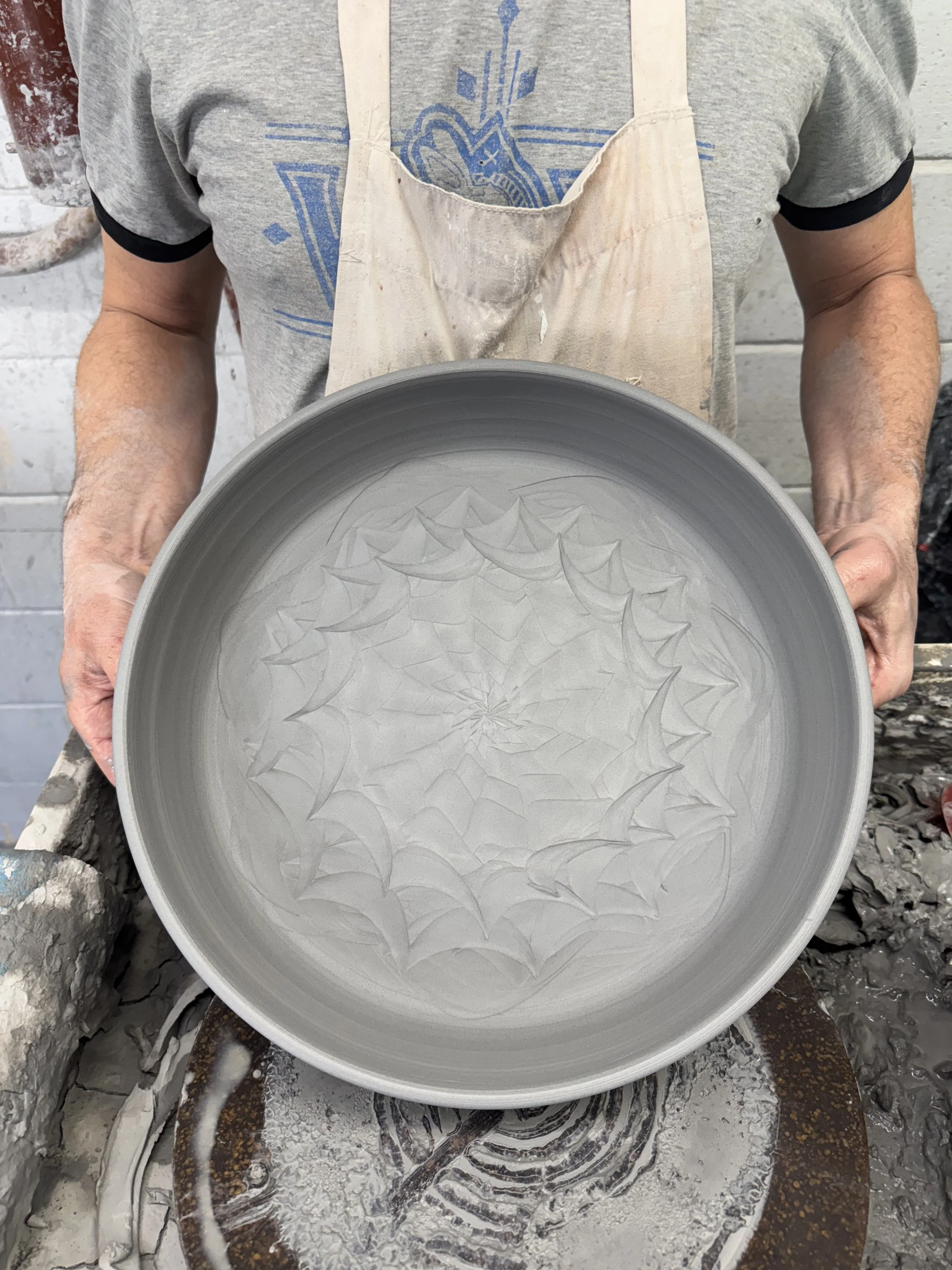 A person holding a large gray ceramic bowl with a web-like carved pattern inside, in a pottery studio.