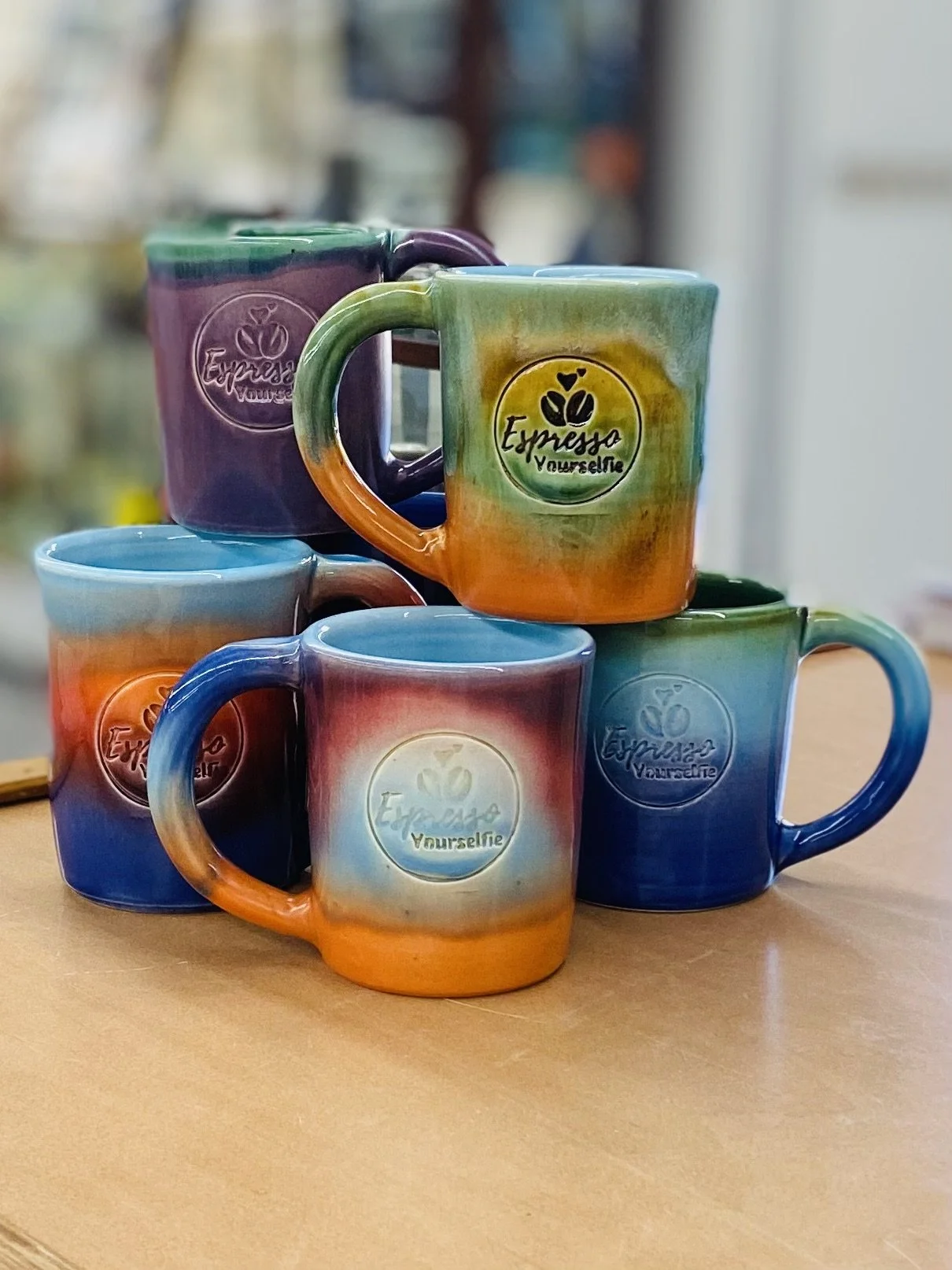 Colorful ceramic mugs with a rainbow pattern and the words "Espresso Yourself" and "Yourselife" embossed on them, stacked on a wooden surface.