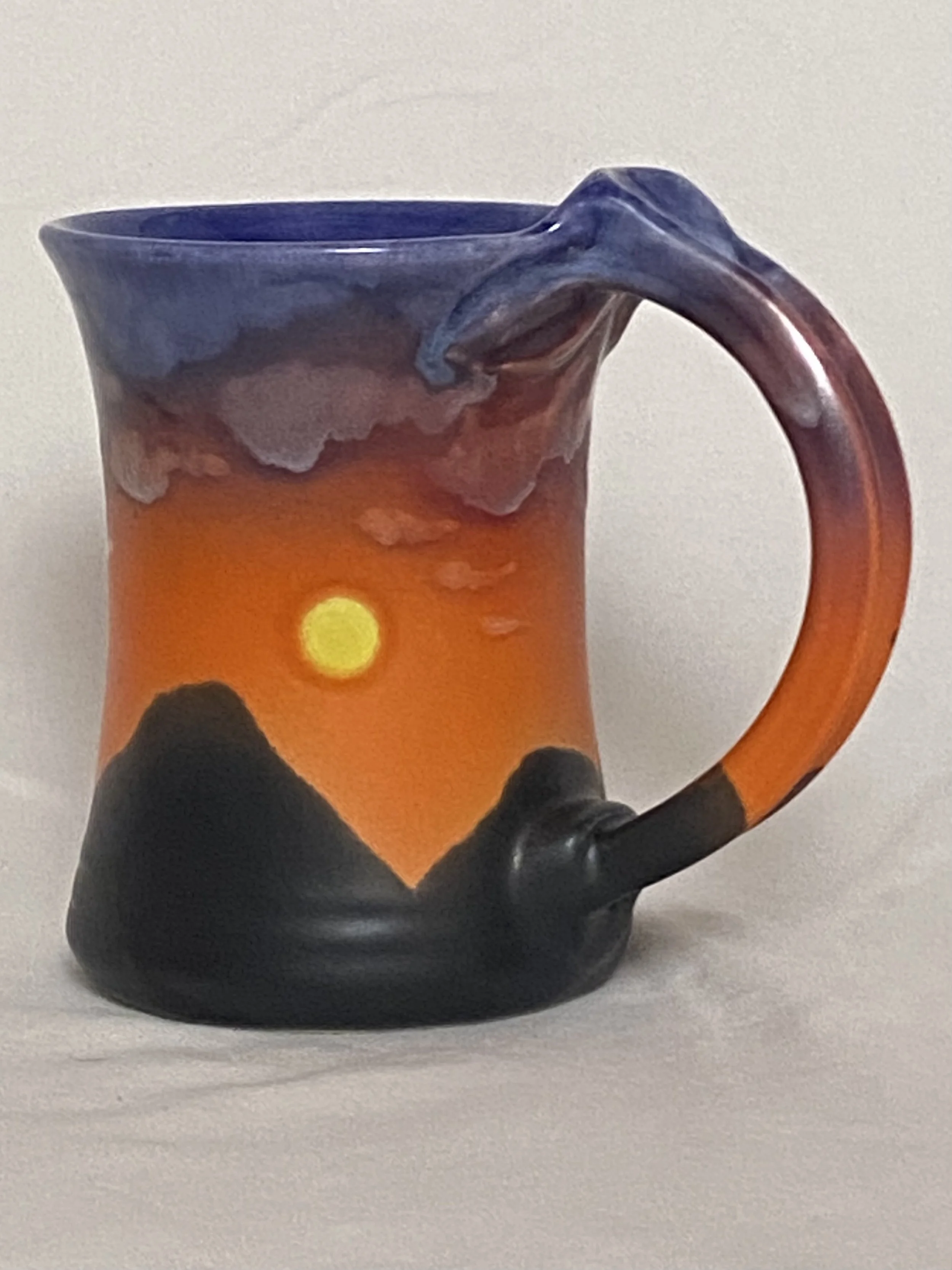 Ceramic coffee mug with a painted sunset scene, featuring the sun setting behind mountains with colorful sky and clouds.