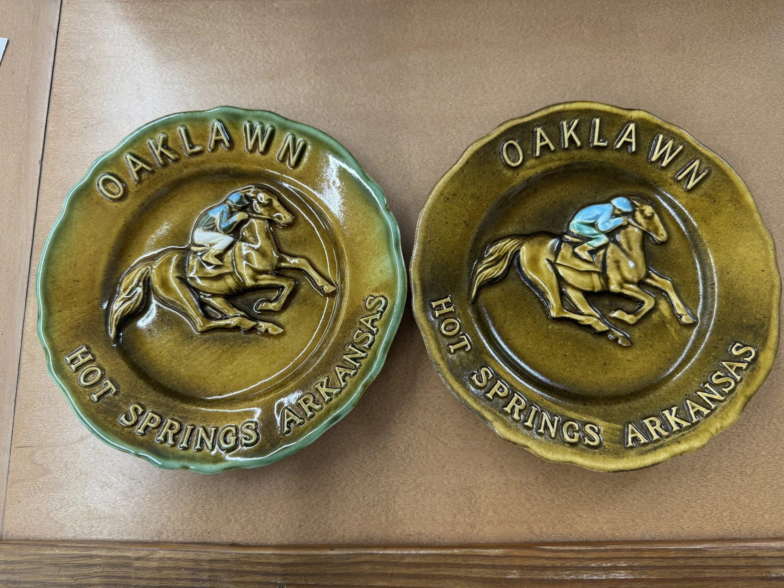 Two decorative ceramic ashtrays with the words "OAKLAWN HOT SPRINGS ARKANSAS" and a horse and rider design, one with a greenish border and the other with a yellowish border, placed on a wooden surface.