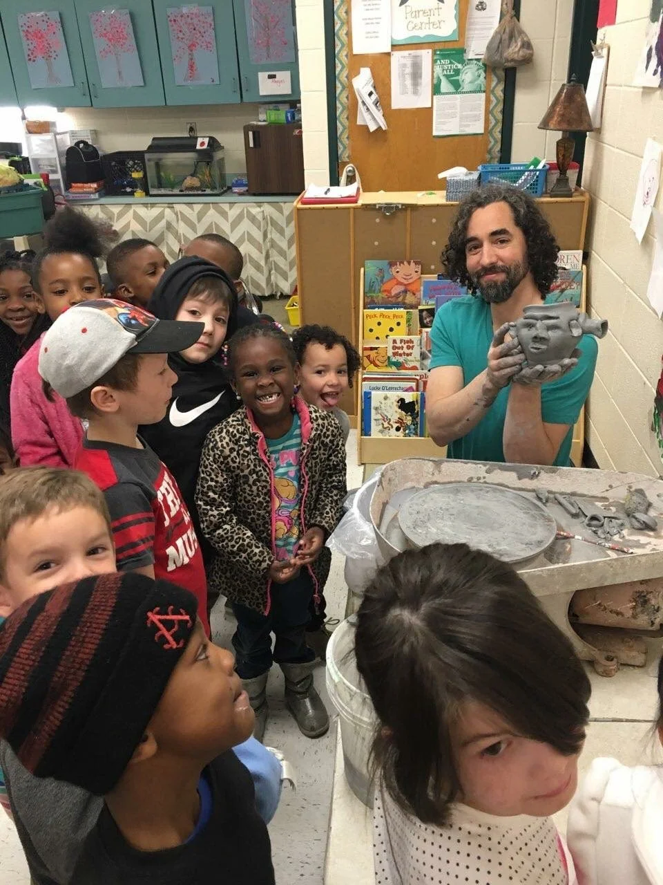 A man showing a ceramic sculpture to a group of children in a classroom or art room.