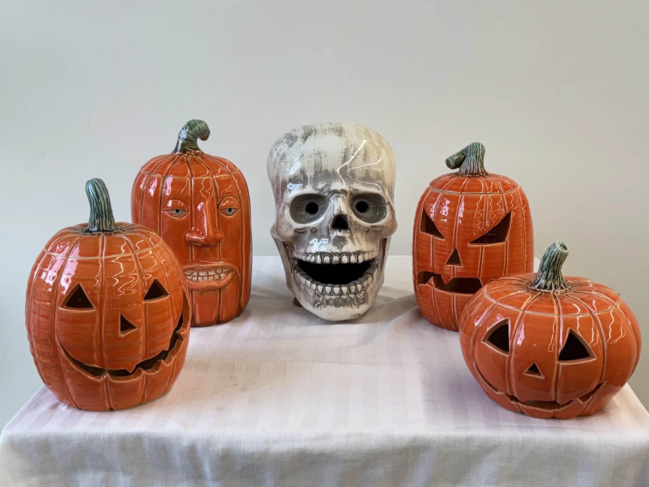 Five Halloween-themed ceramic decorations on a table, including three pumpkins with carved faces, one with a face design, and a human skull.