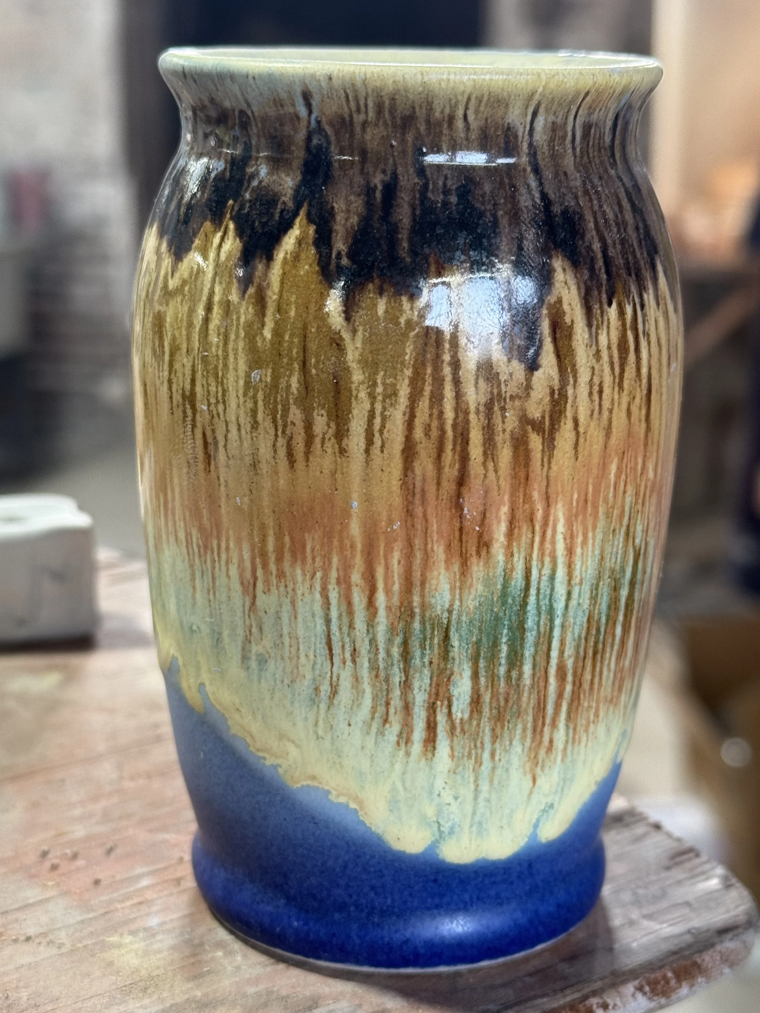 A colorful ceramic vase with a glossy finish, featuring a gradient glaze that transitions from dark brown and black at the top, through shades of orange and yellow, to green and blue at the bottom.